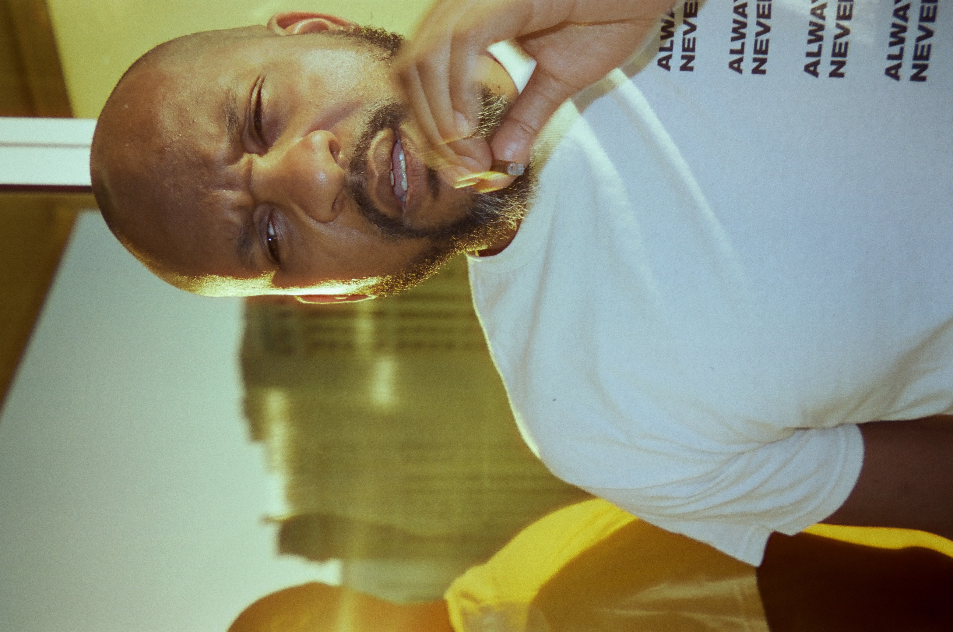 A bald, bearded man laughs with his mouth open, one hand raised near his face, wearing a white shirt printed with bold black text.