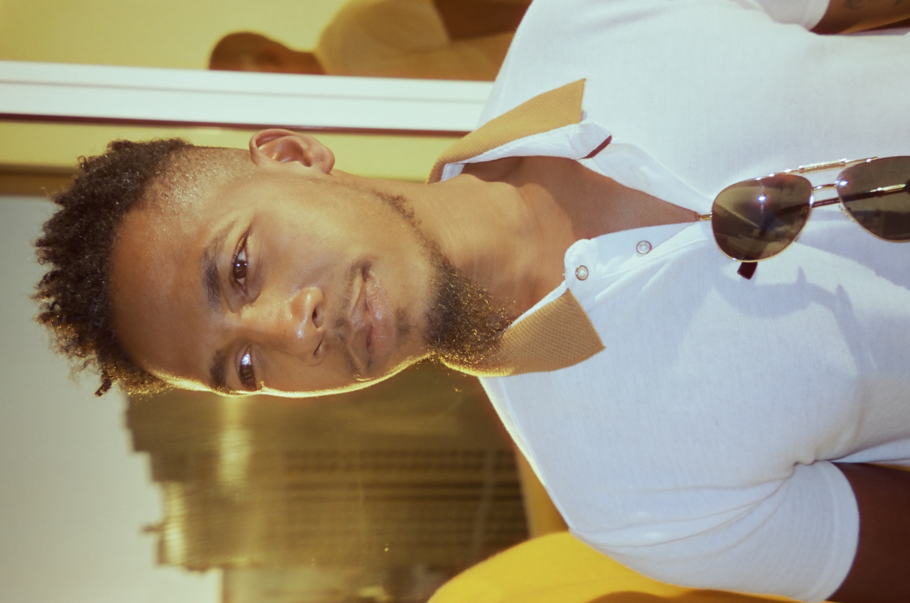 A young man with short curly hair and a light beard looks past the camera, aviator sunglasses hanging from the collar of his white shirt.