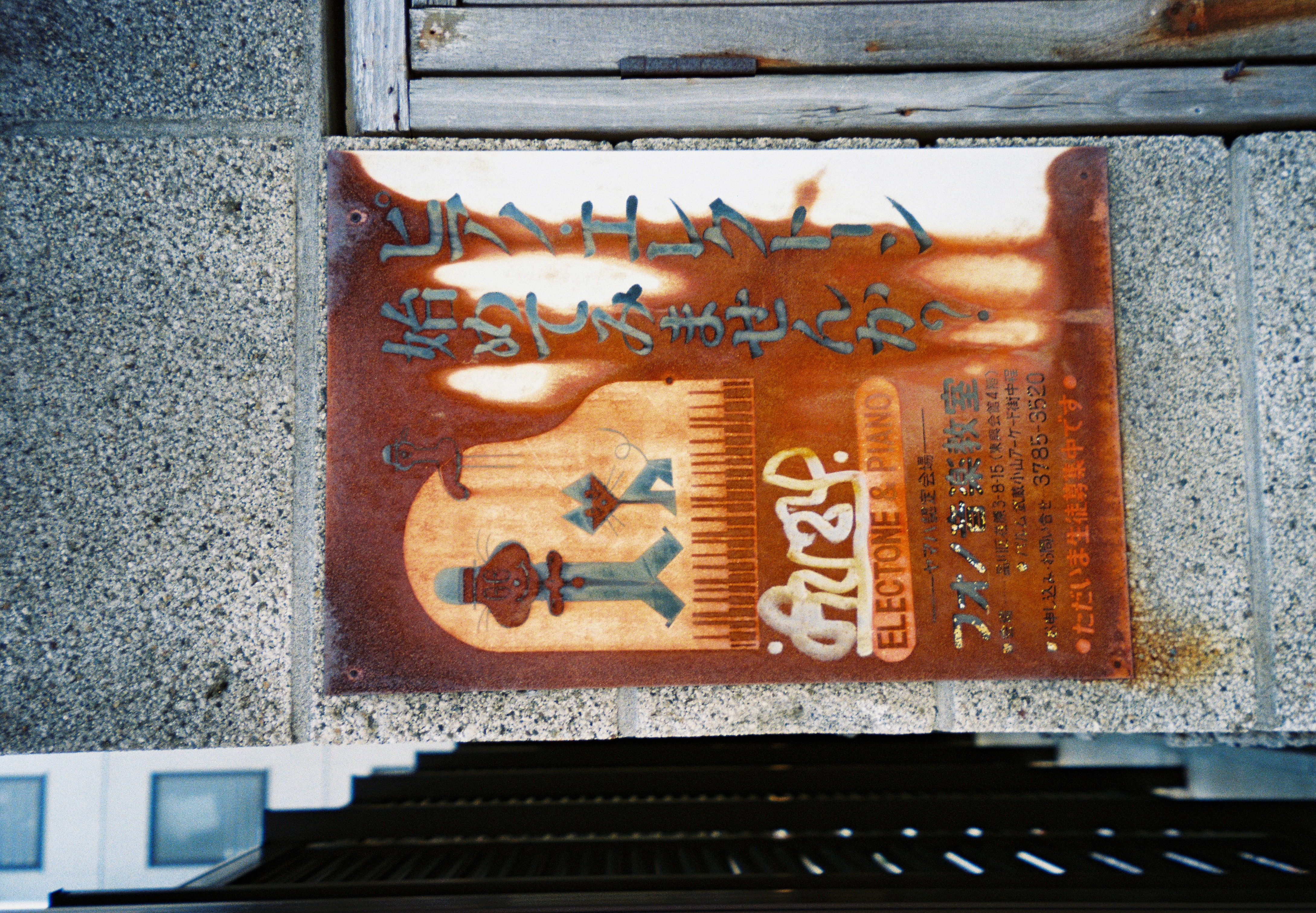 A rusted enamel sign bolted to a granite pillar reads 'ELECTONE & PIANO' above illustrated cat characters and a keyboard, with Japanese text and a phone number.