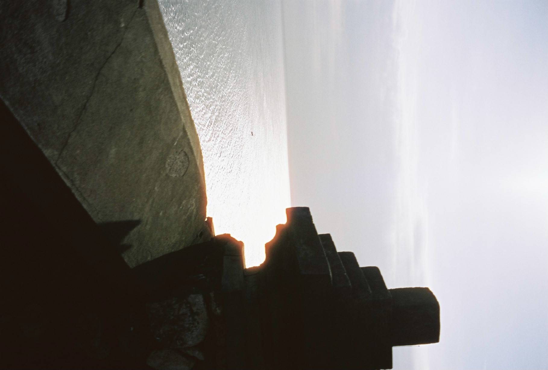 A dark stepped structure occupies the right edge above a shadowed sloping paved surface and a broad expanse of reflective water.