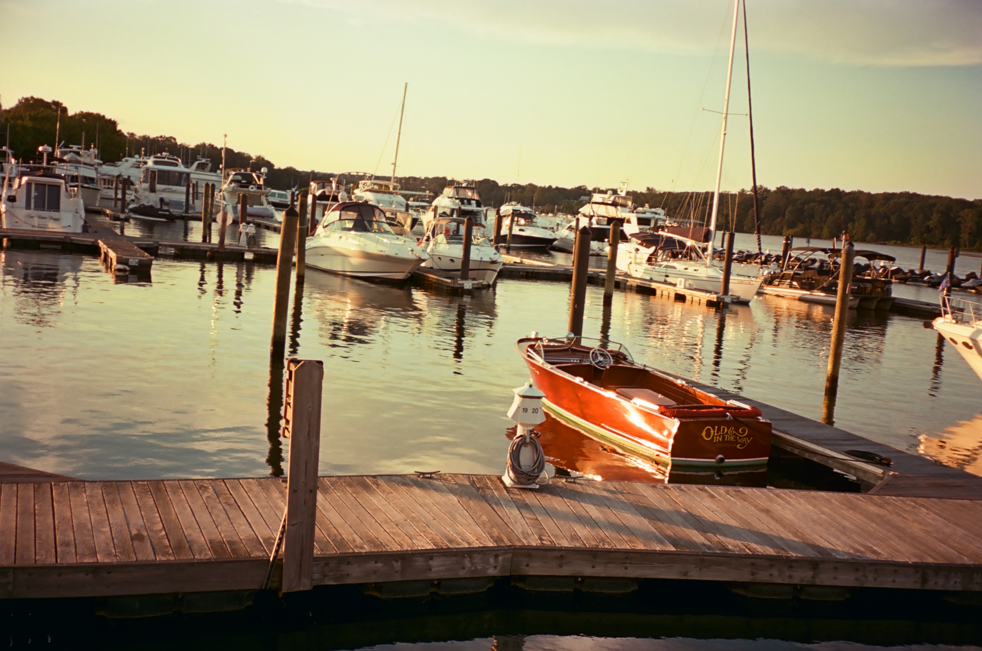 Wooden runabout lettered Old & In the Way moored beside a plank dock, with white powerboats, two sailboat masts, and calm water in warm low light.