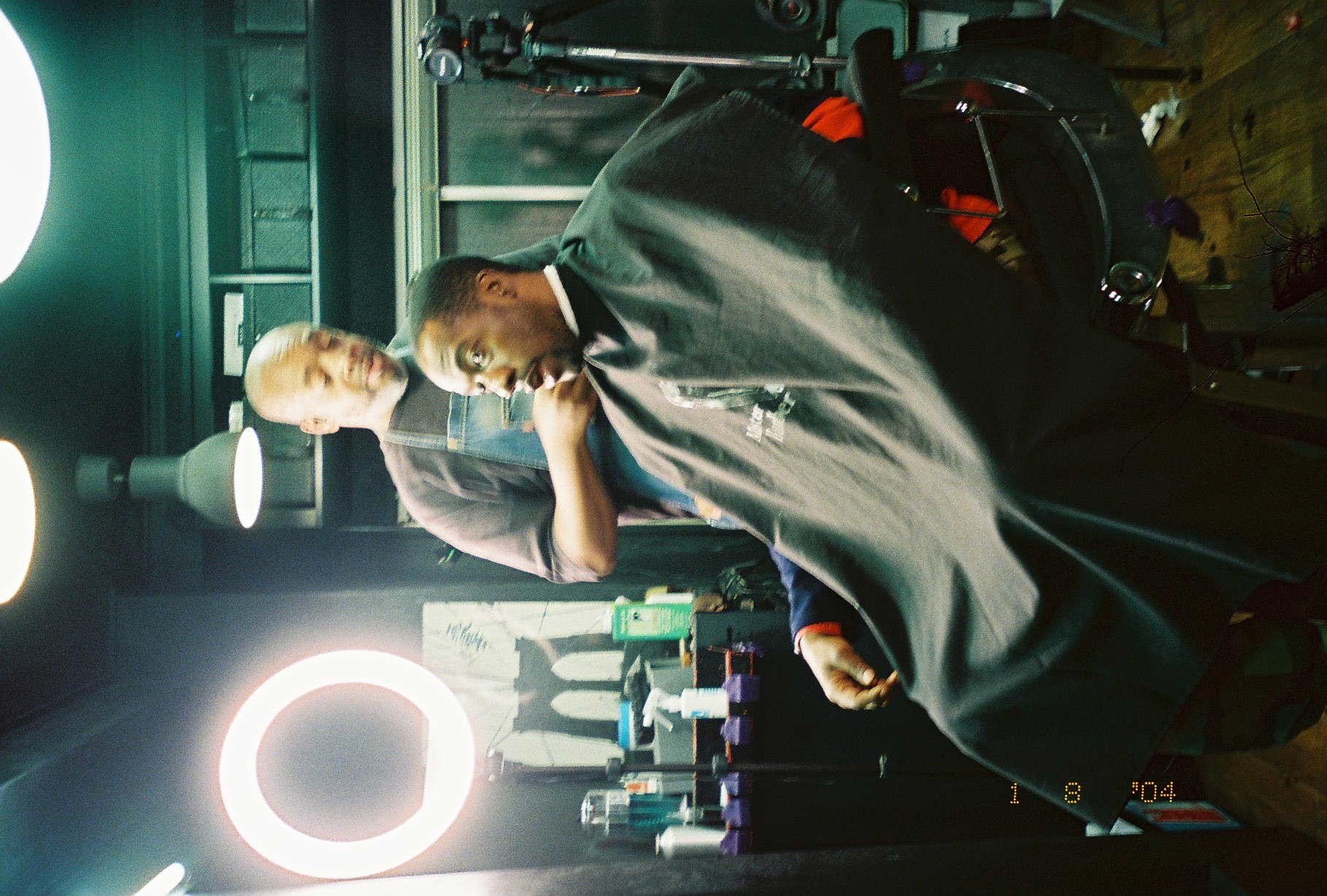 A barber in a denim apron trims a seated, caped client's beard beside a glowing circular ring light in a dim shop.