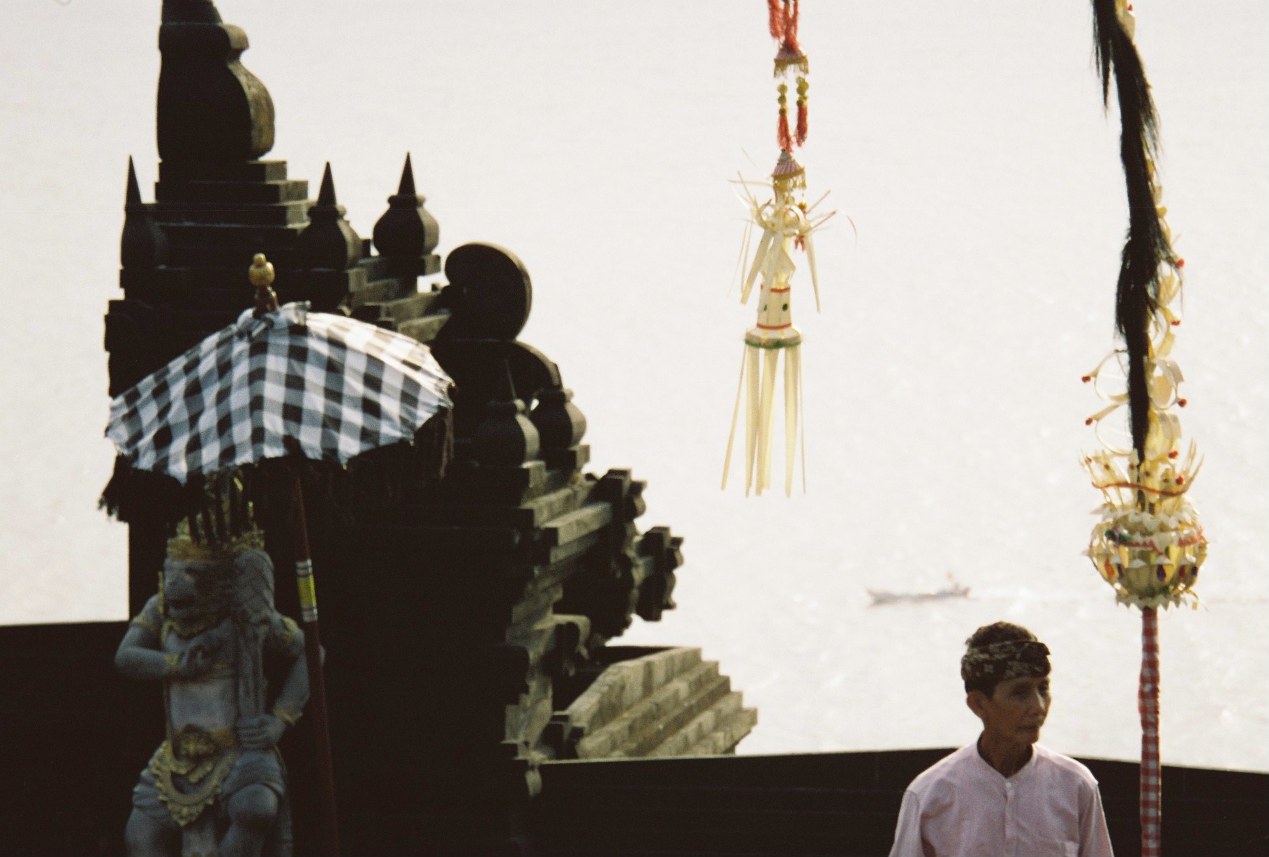 A man in a white shirt and patterned headband stands before a black stone temple roof, a checkered umbrella, a carved guardian statue, and hanging tasseled ornaments.