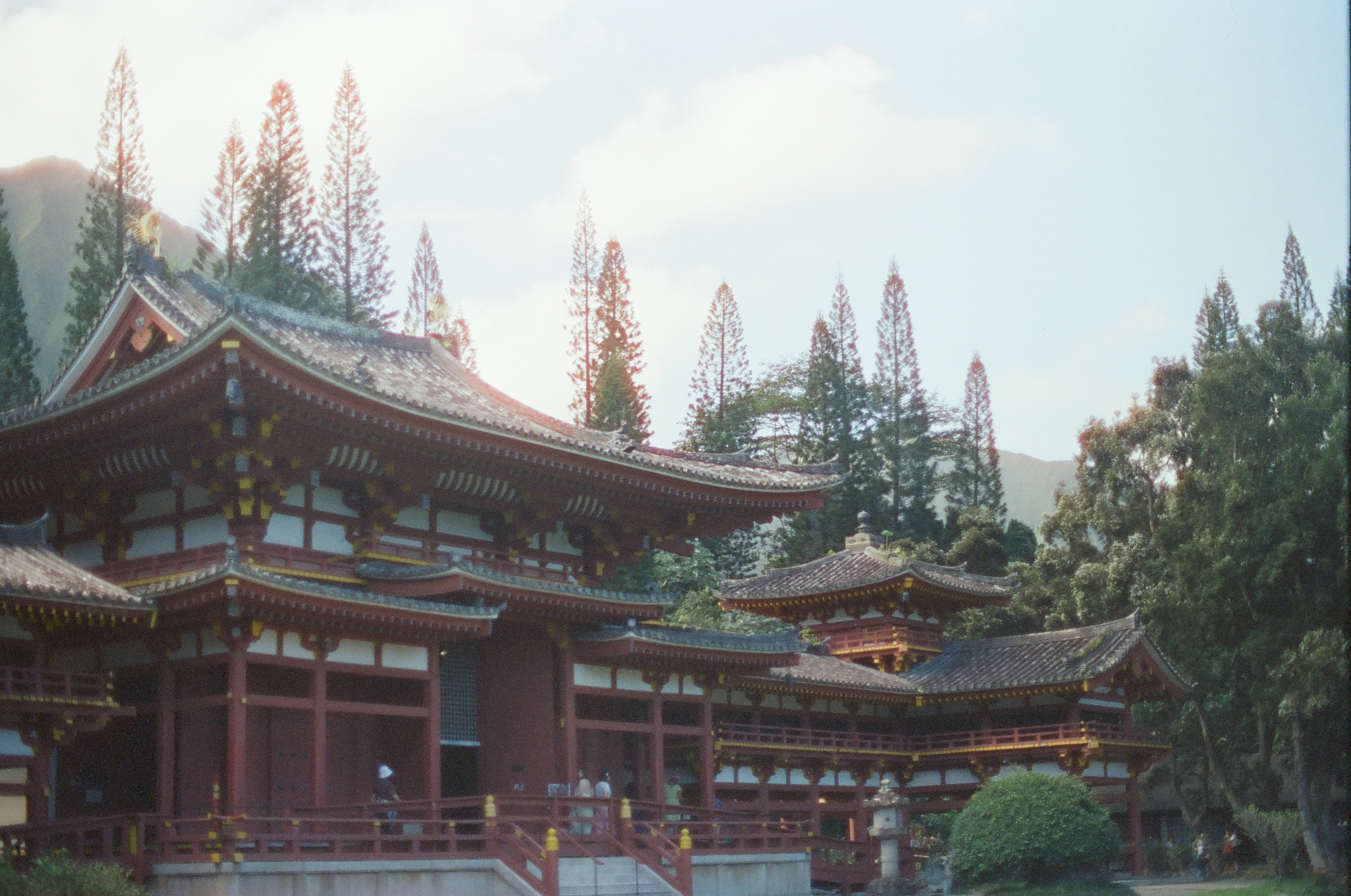A red and white multi-tiered temple with dark tiled roofs rises among tall pine trees, with hazy green mountains and small figures crossing a railed bridge below.