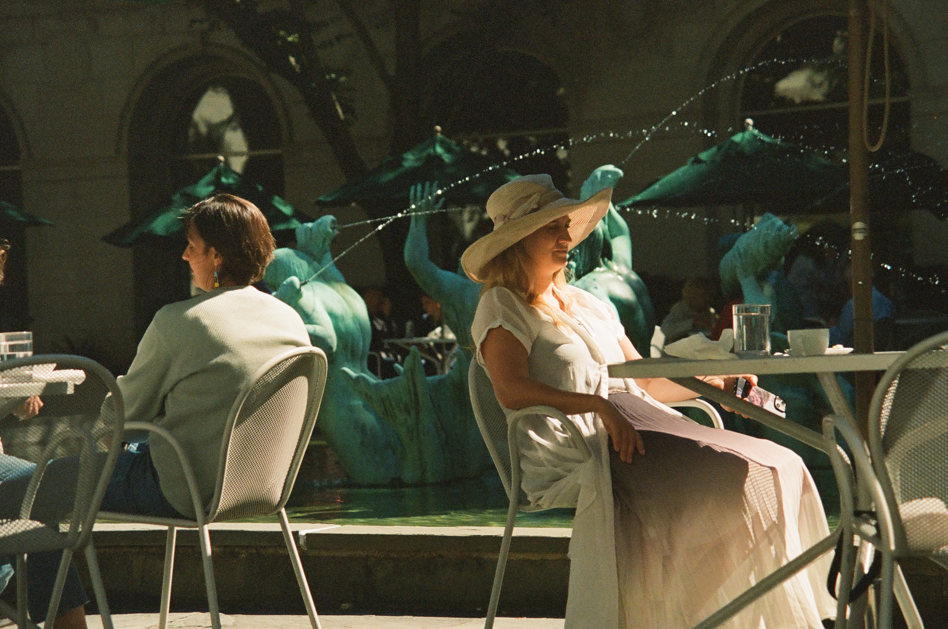 Two people sit at terrace tables before green animal sculptures and a shadowed arched entrance.