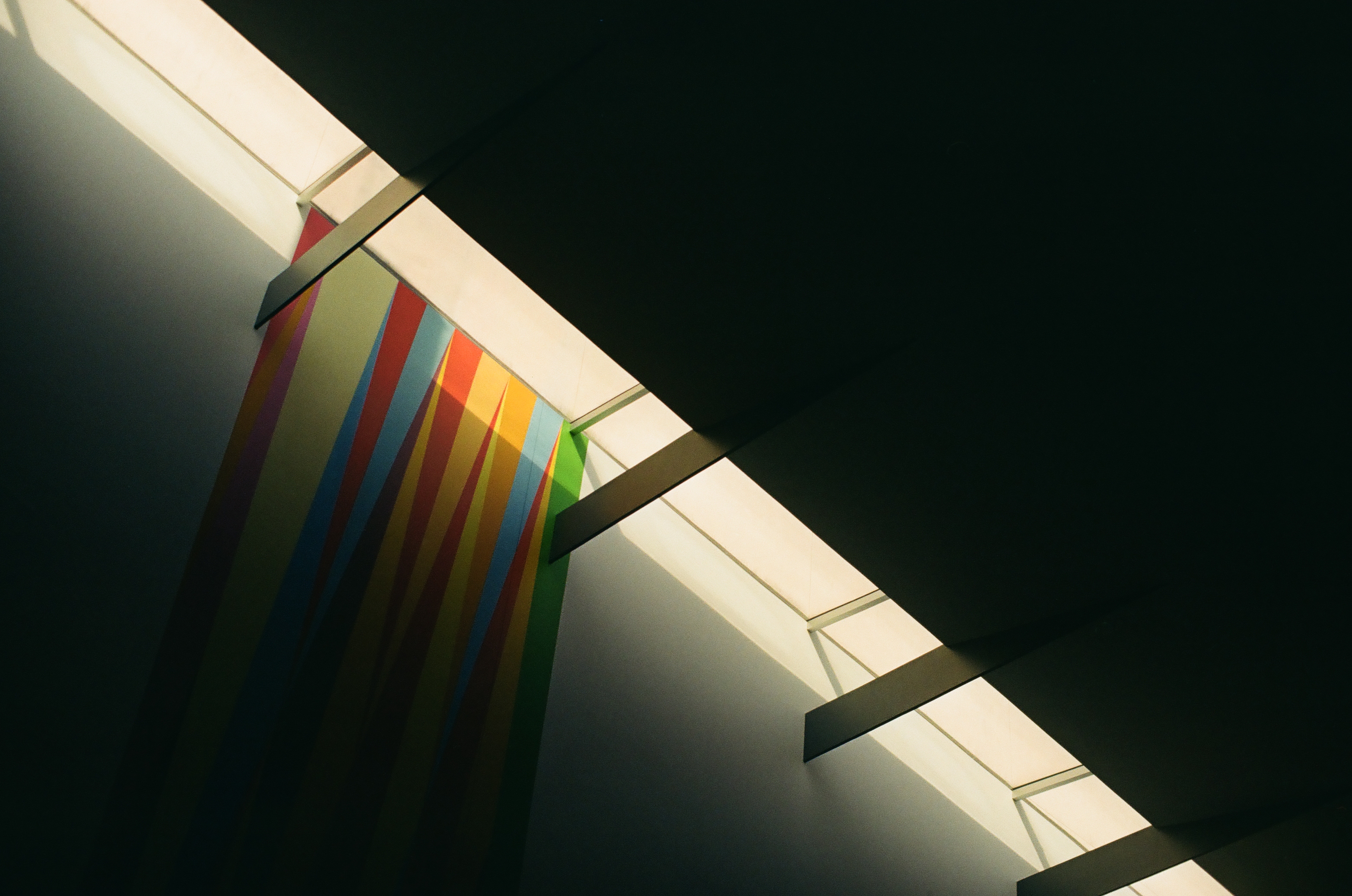 A vertically striped hanging in red, orange, yellow, blue, green, and pink tapers to points beneath a diagonal strip of lit ceiling panels.