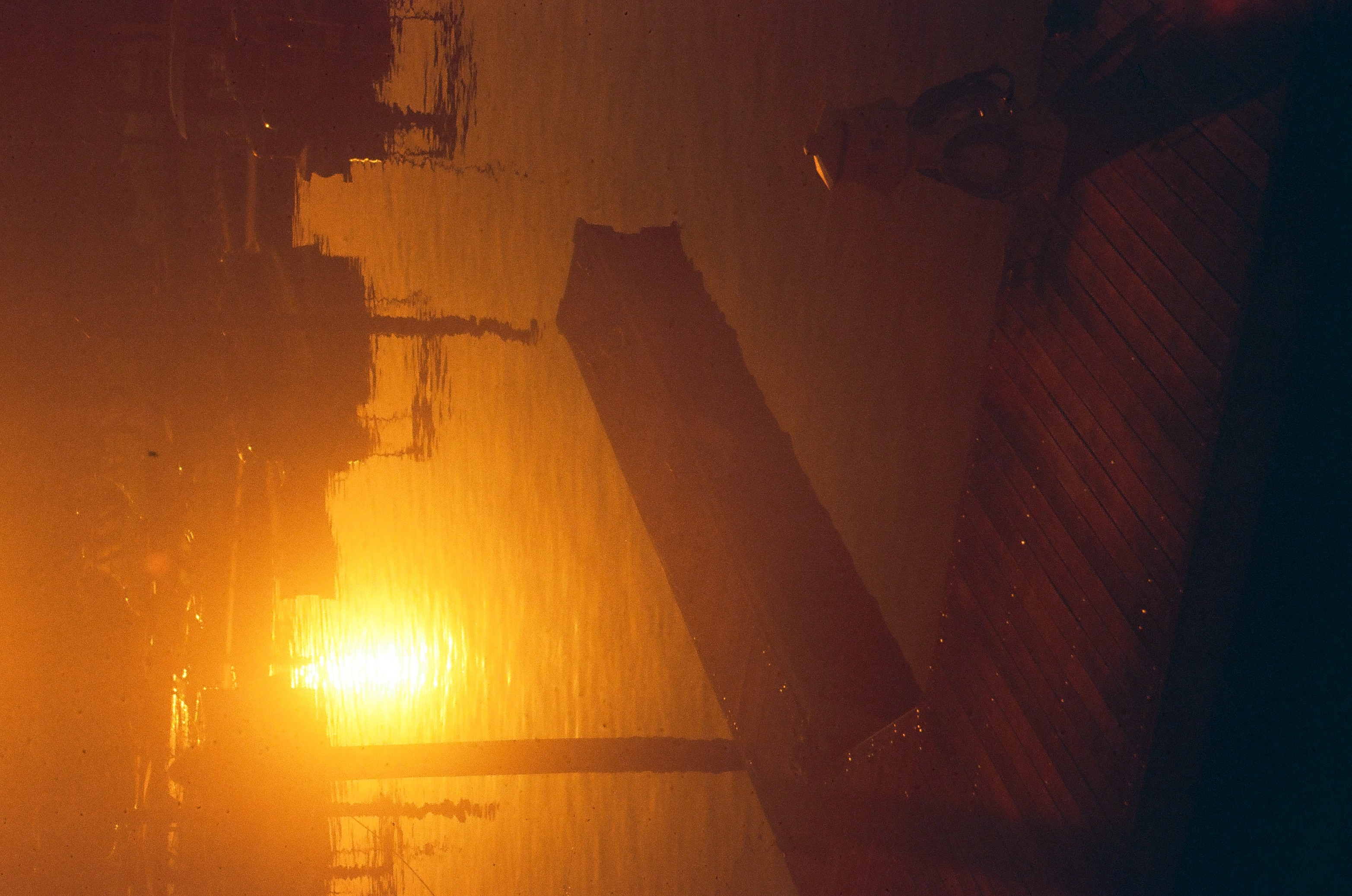 A bright orange light glows behind dark angular structures and a slanted pole, its warm color bleeding through heavy film grain.