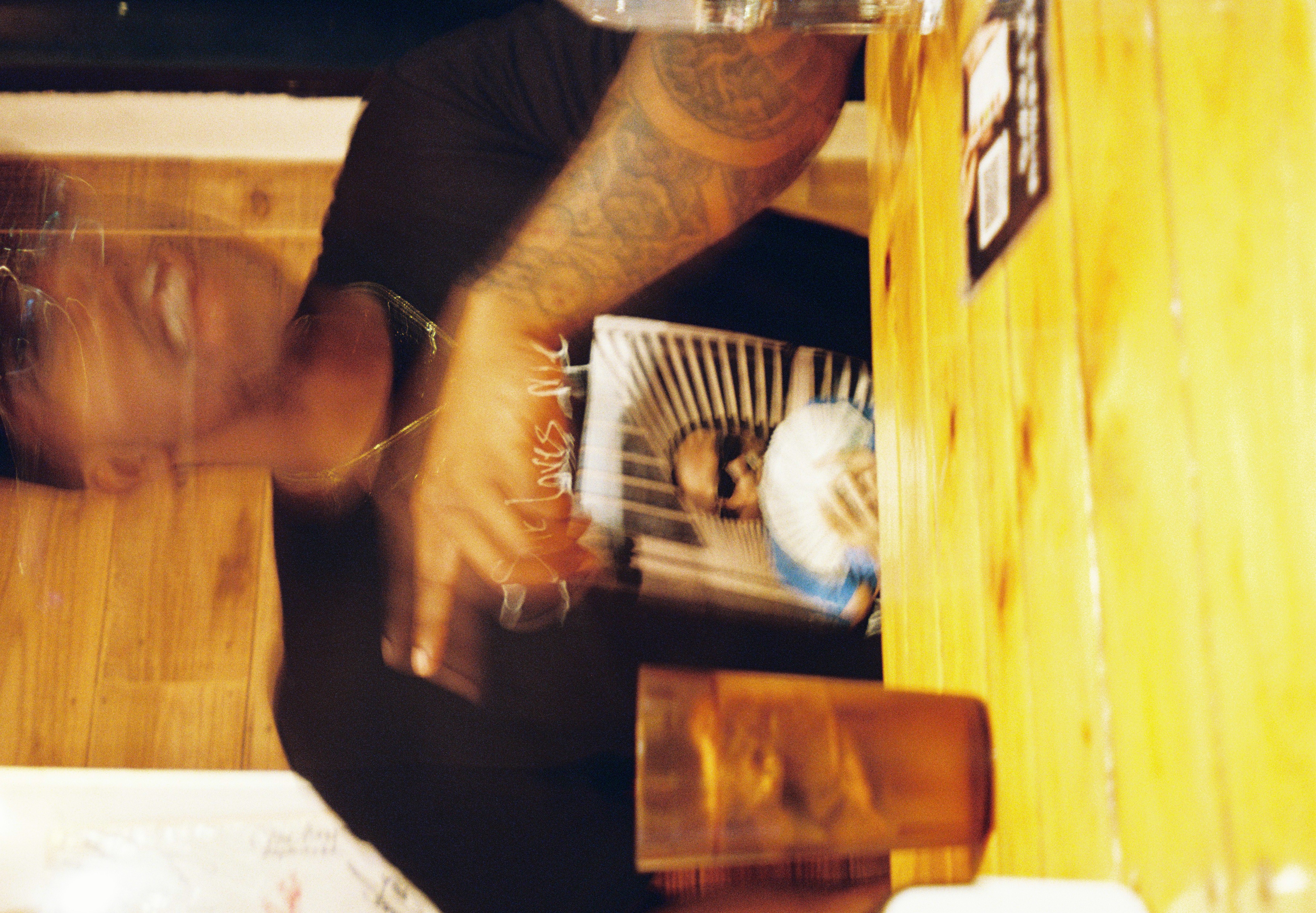 A person's blurred gesture crosses a pale wooden table, a glass of iced drink standing sharp and still beside them.