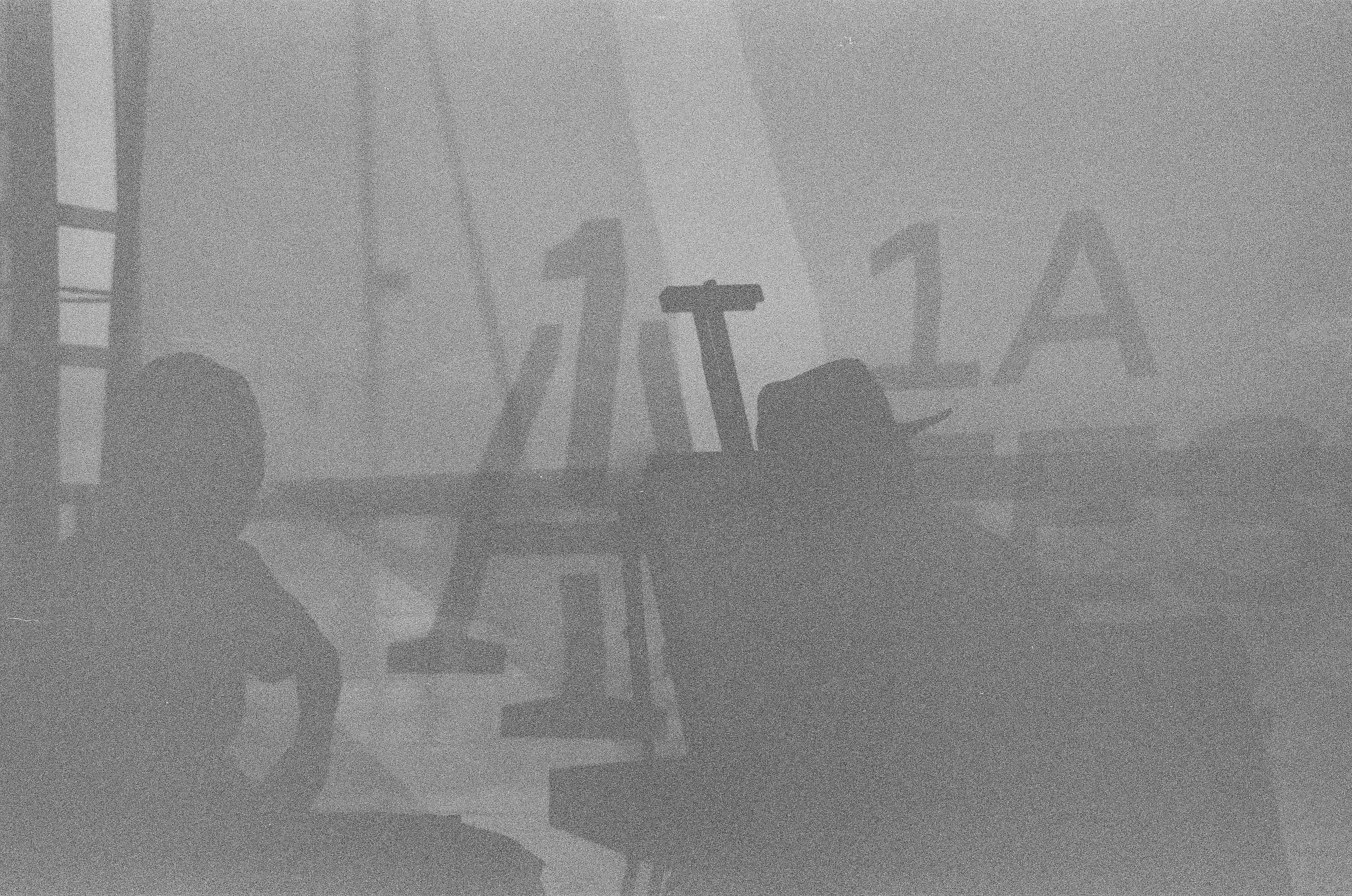 A pale monochrome scene shows a seated silhouette, large cropped letters, and faint aircraft or terminal structures through heavy haze.