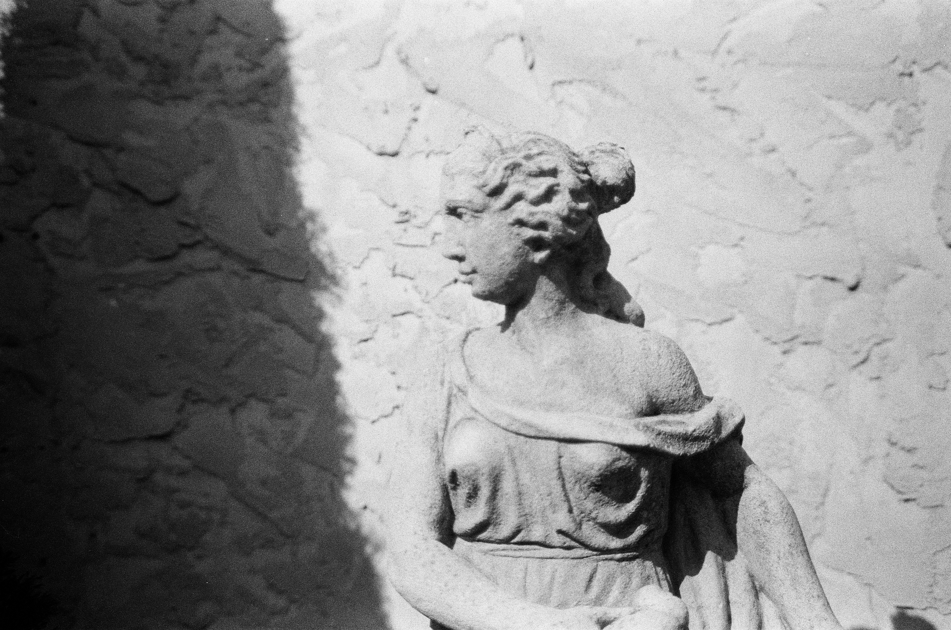 A weathered stone female figure stands against a rough wall sharply divided by sunlight and shadow.