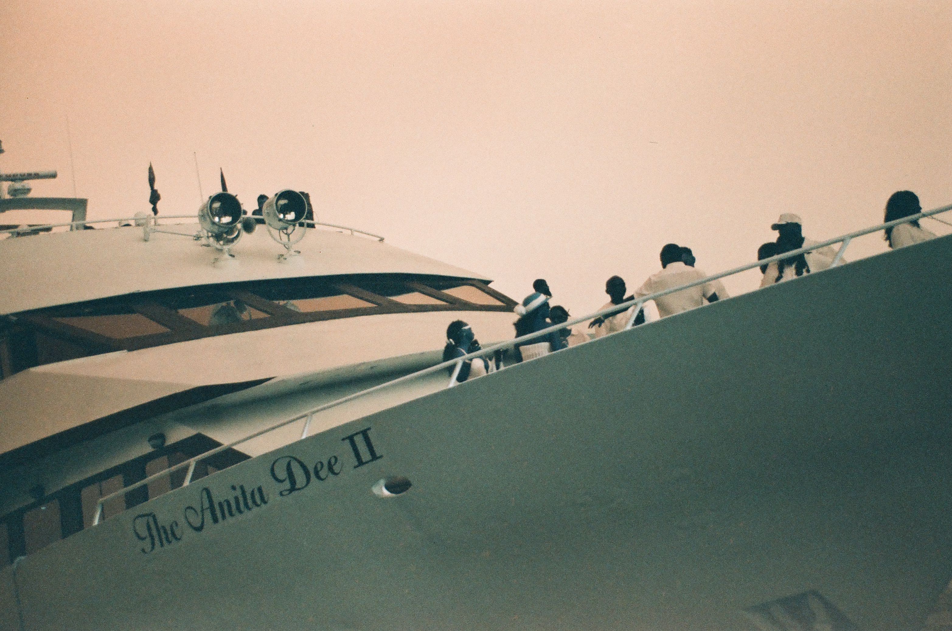 Passengers lean along the railing of a yacht named The Anita Dee II, its radar mast and spotlights outlined against a peach evening sky.