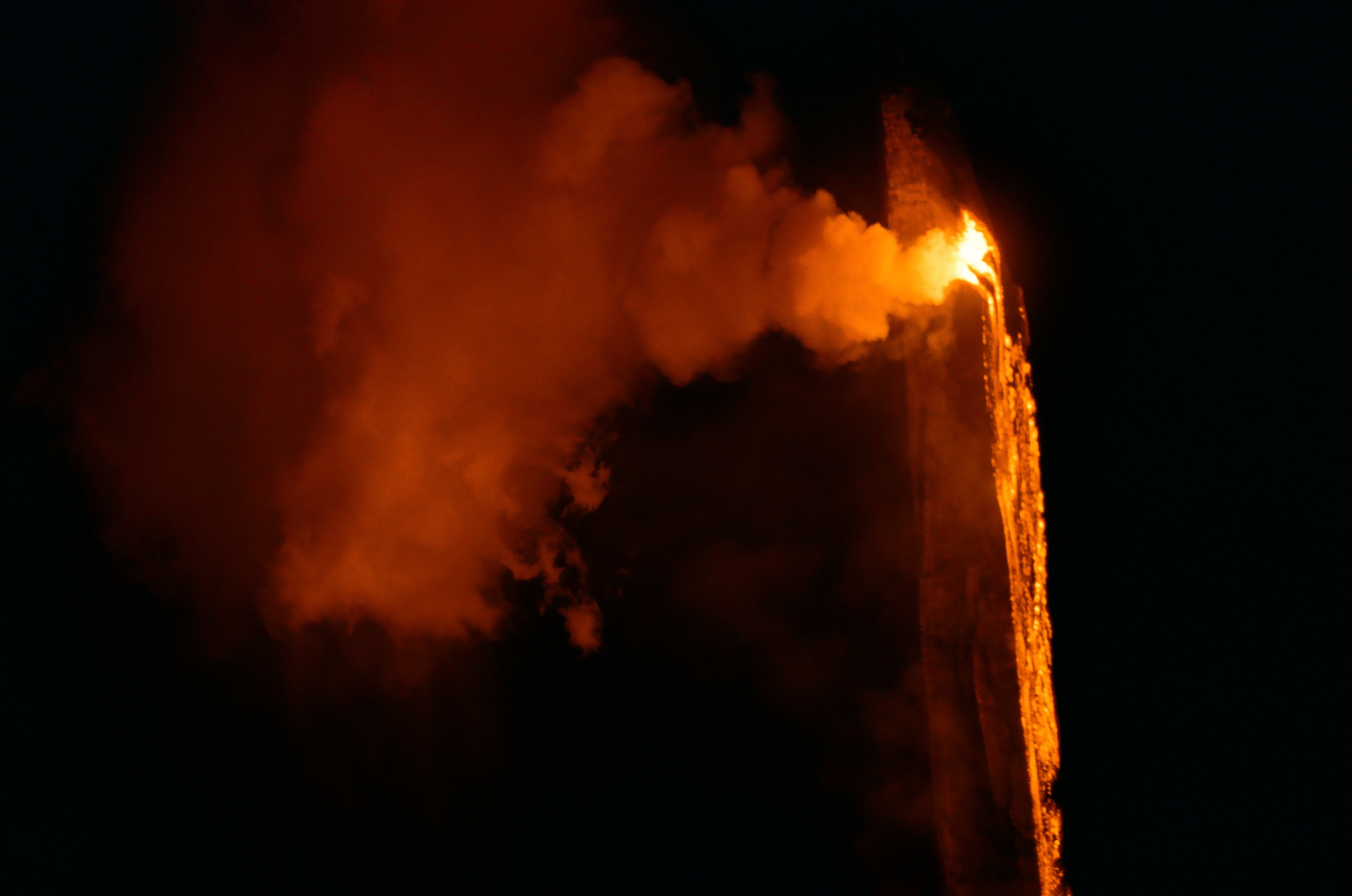 A vertical stream of orange lava spills down a dark edge beneath thick glowing smoke at night.