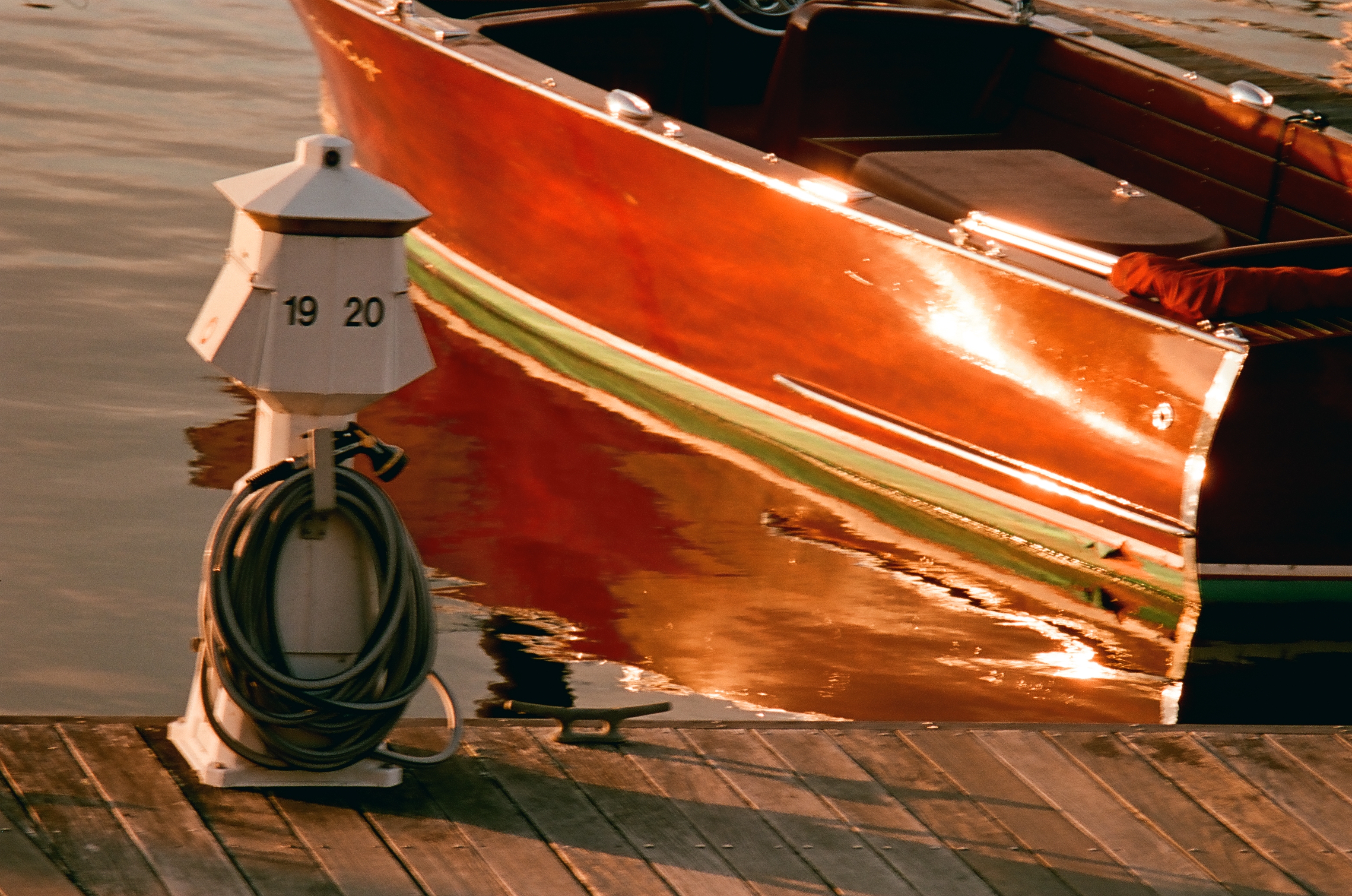 Varnished wooden boat hull reflecting low orange sunlight beside a white dock post numbered 19 and 20 with a coiled gray hose on weathered planks.