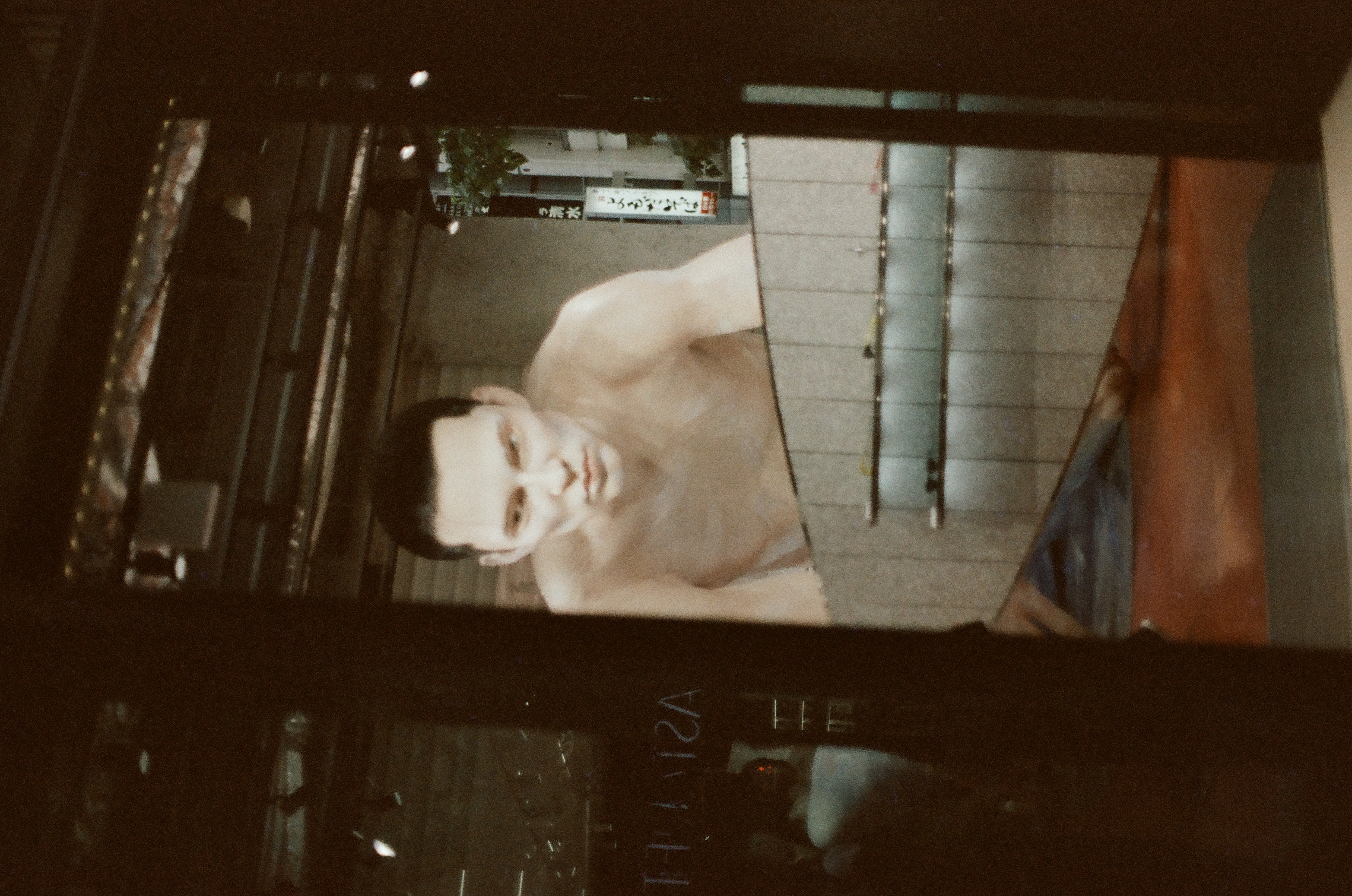 A large figure image, showing a shirtless man leaning over a curved tiled counter, fills a lit storefront window seen through a dark open doorway.