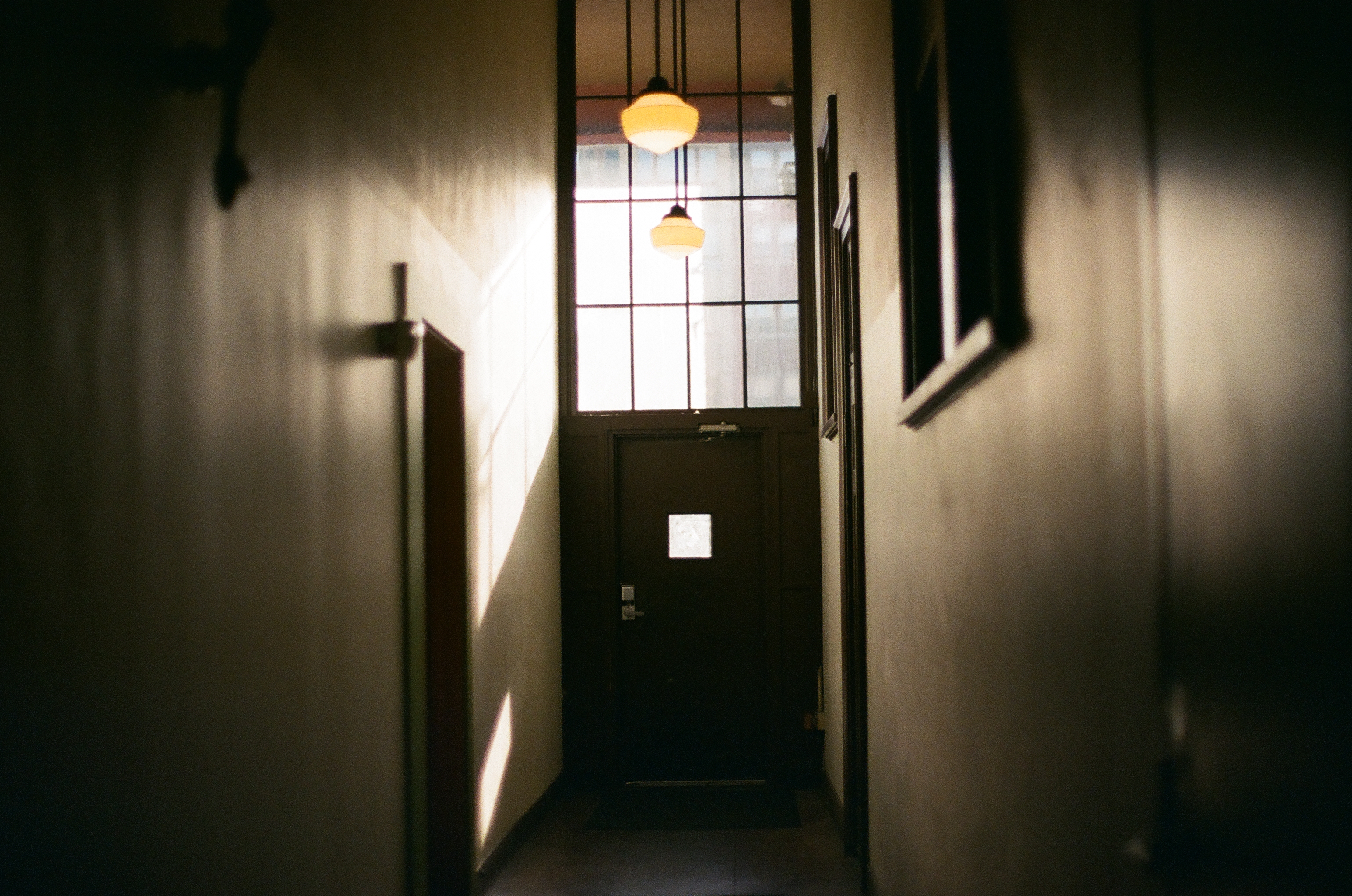Two warm pendant lamps lead down a narrow dark hallway toward a closed glass-paneled door.