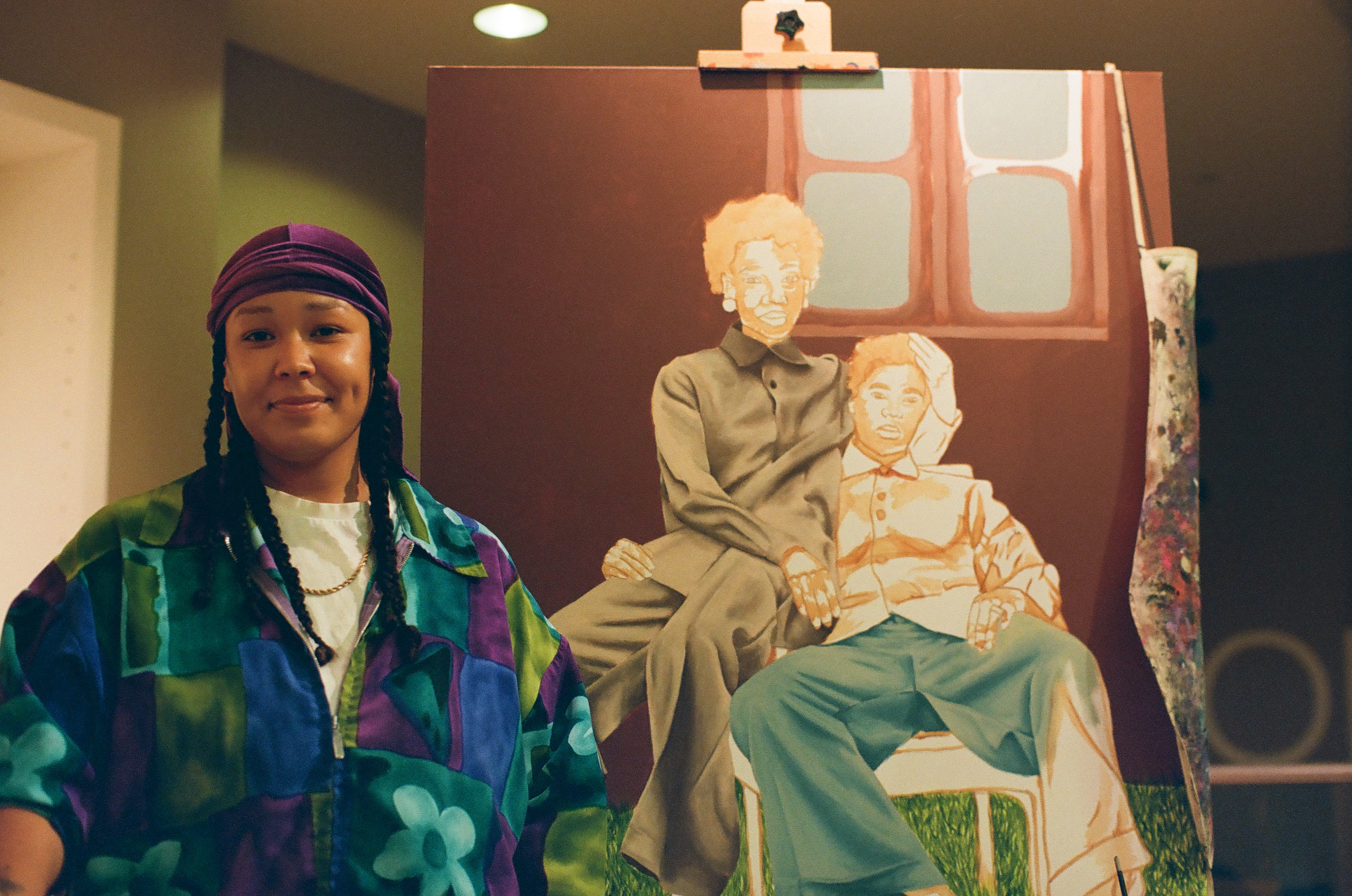 A person faces the camera in a multicolored jacket before a painting of two seated figures.