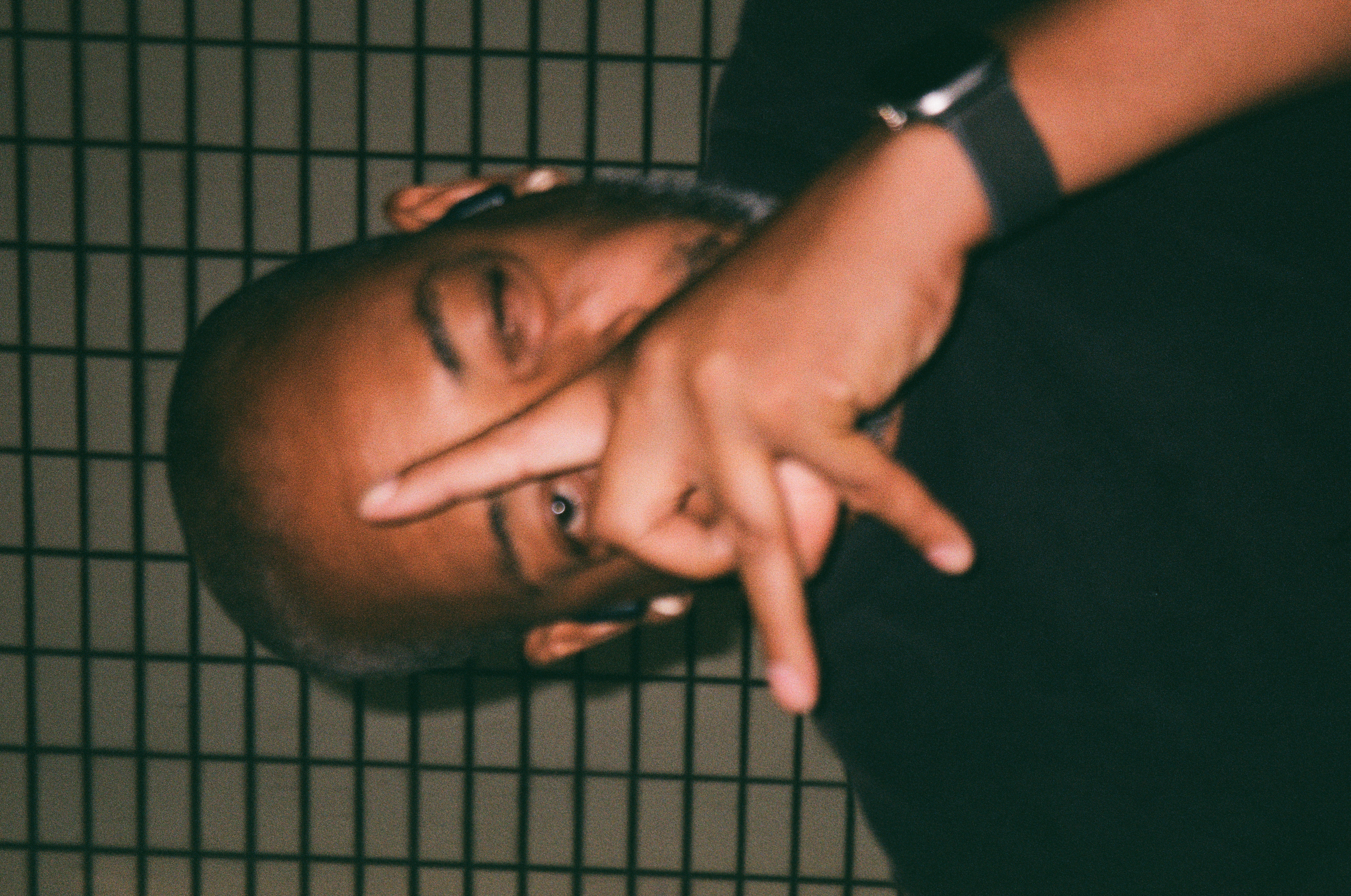 A man with a shaved head and gray beard holds spread fingers across his face, one eye visible between them, before a wall of small rectangular tiles.