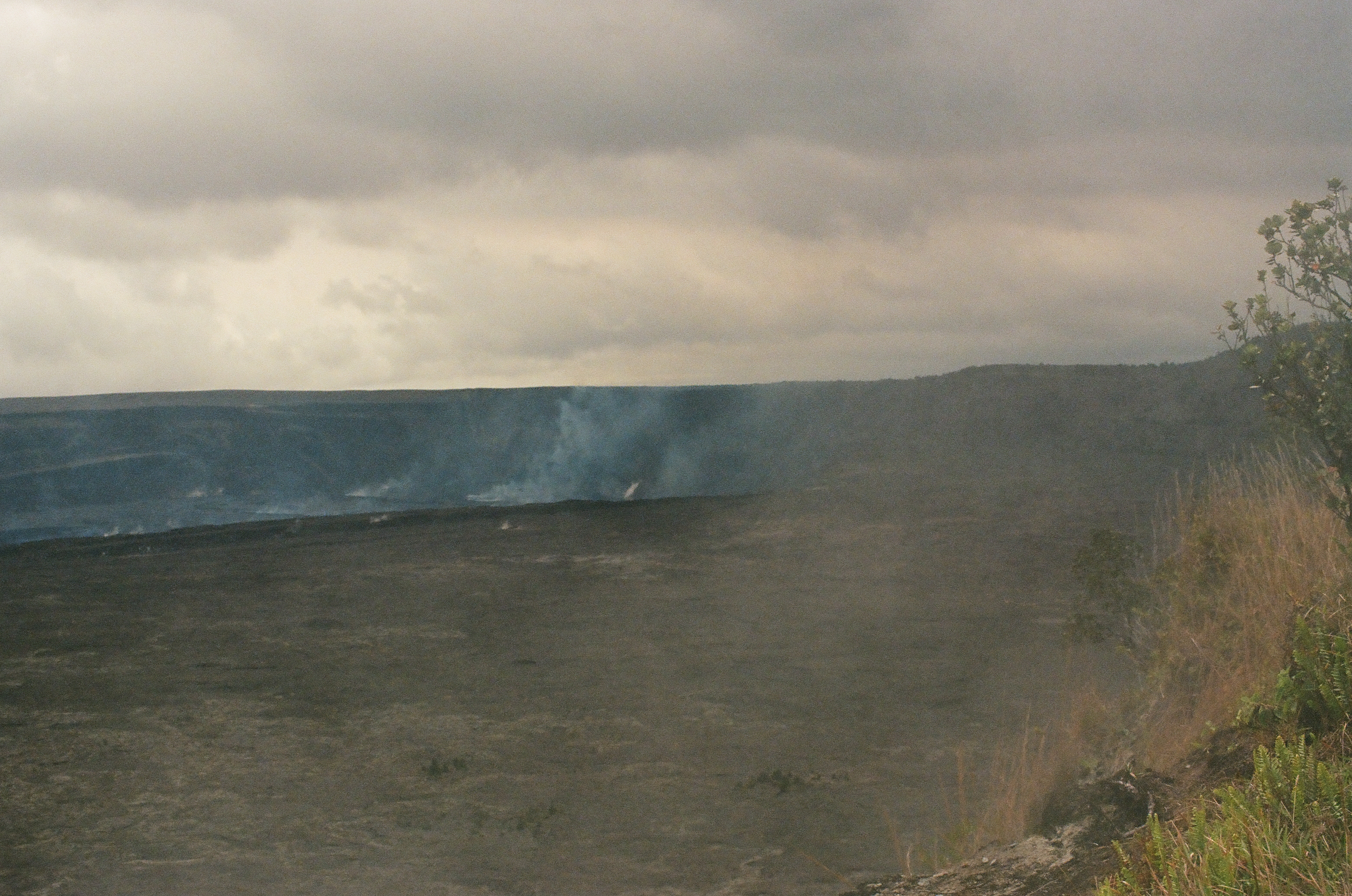 A broad dark crater lies beneath heavy clouds while pale steam rises from several distant points.