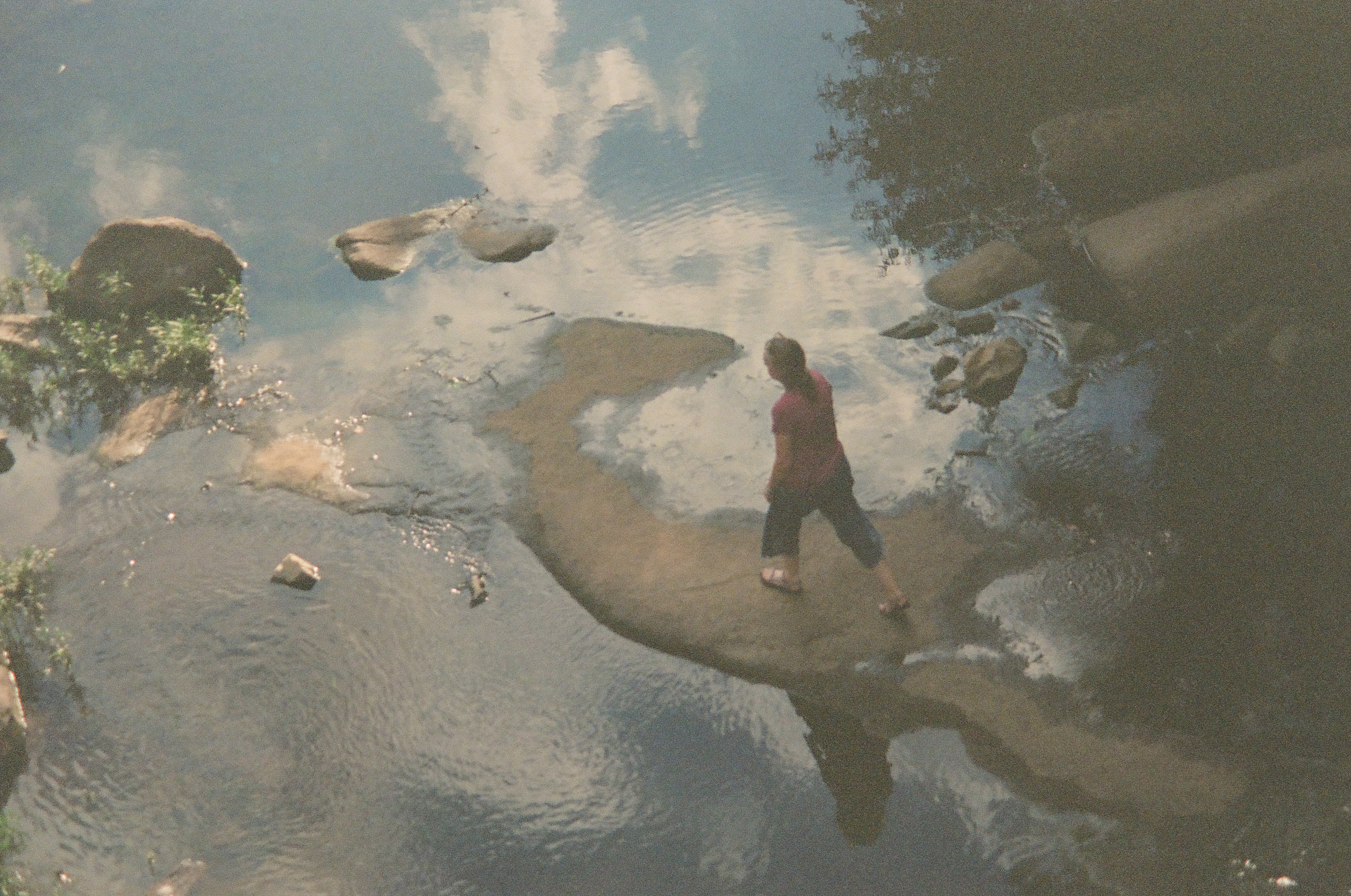 Viewed from above, a person in a red shirt walks across a pale sandbar in shallow water that reflects clouds and surrounding rocks.
