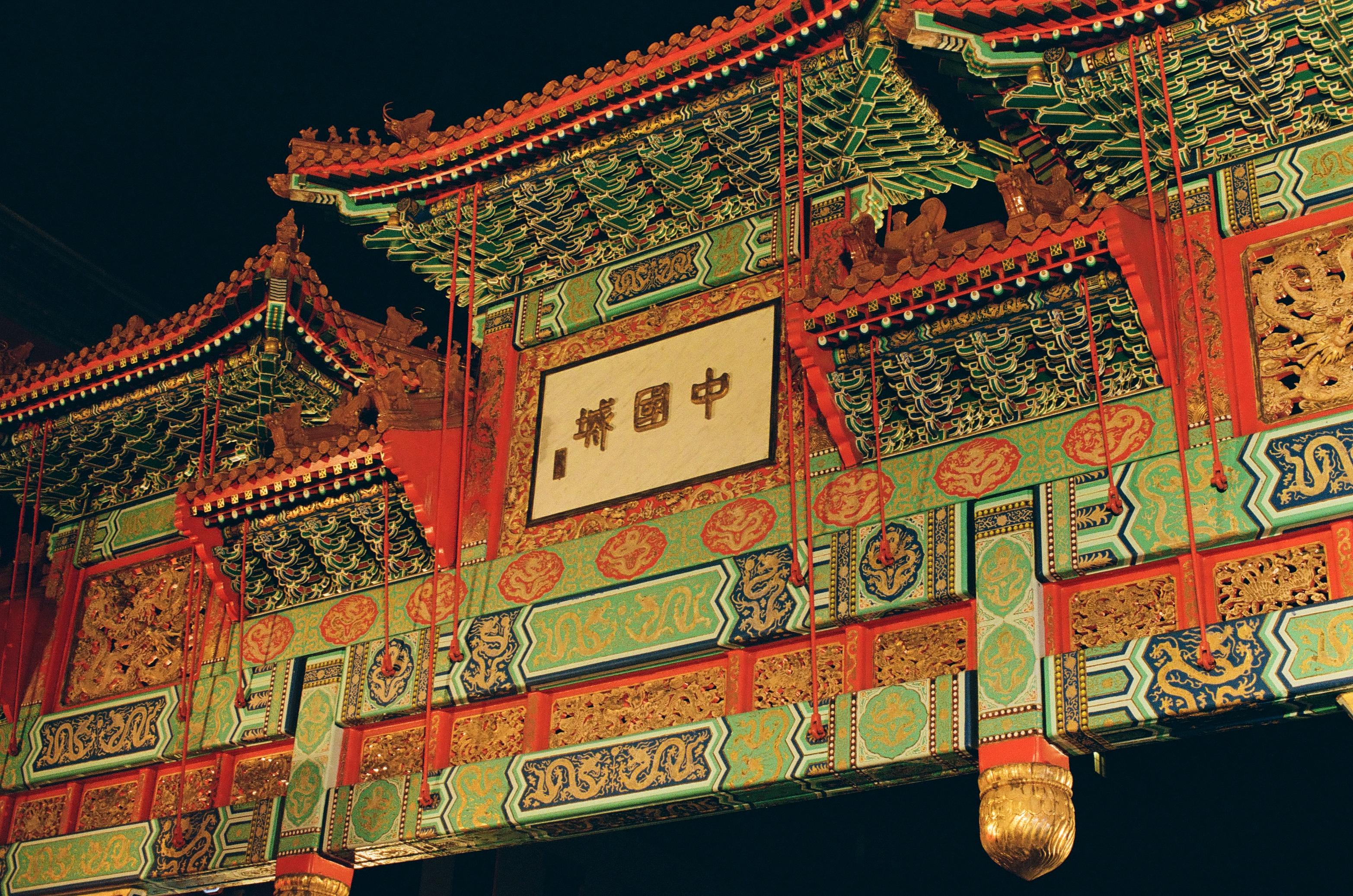A richly painted ceremonial gate with layered roofs and carved brackets glows red, green, and gold against black sky.