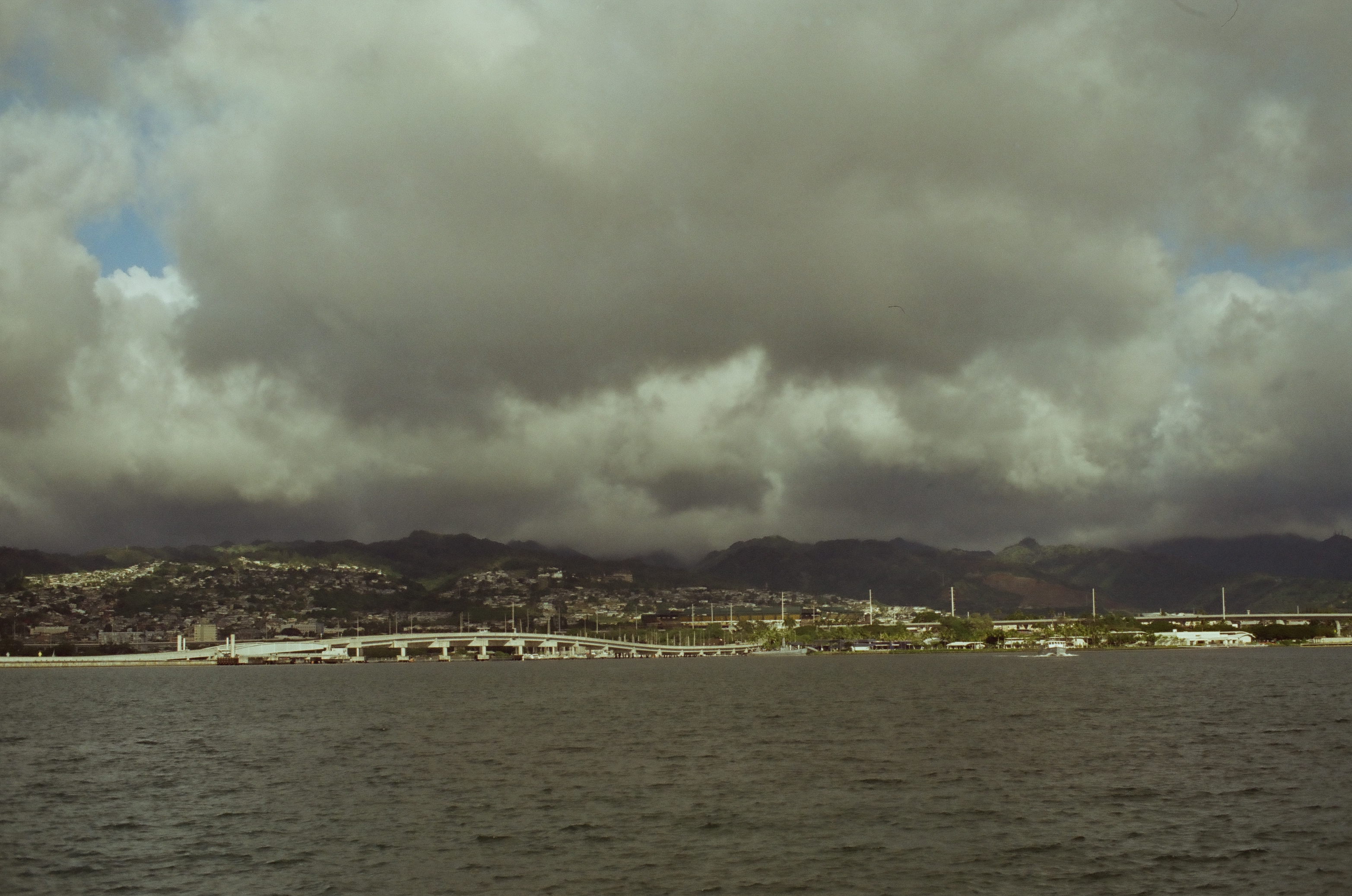 Dark mountains and a low developed shoreline sit beneath thick gray clouds across open water.