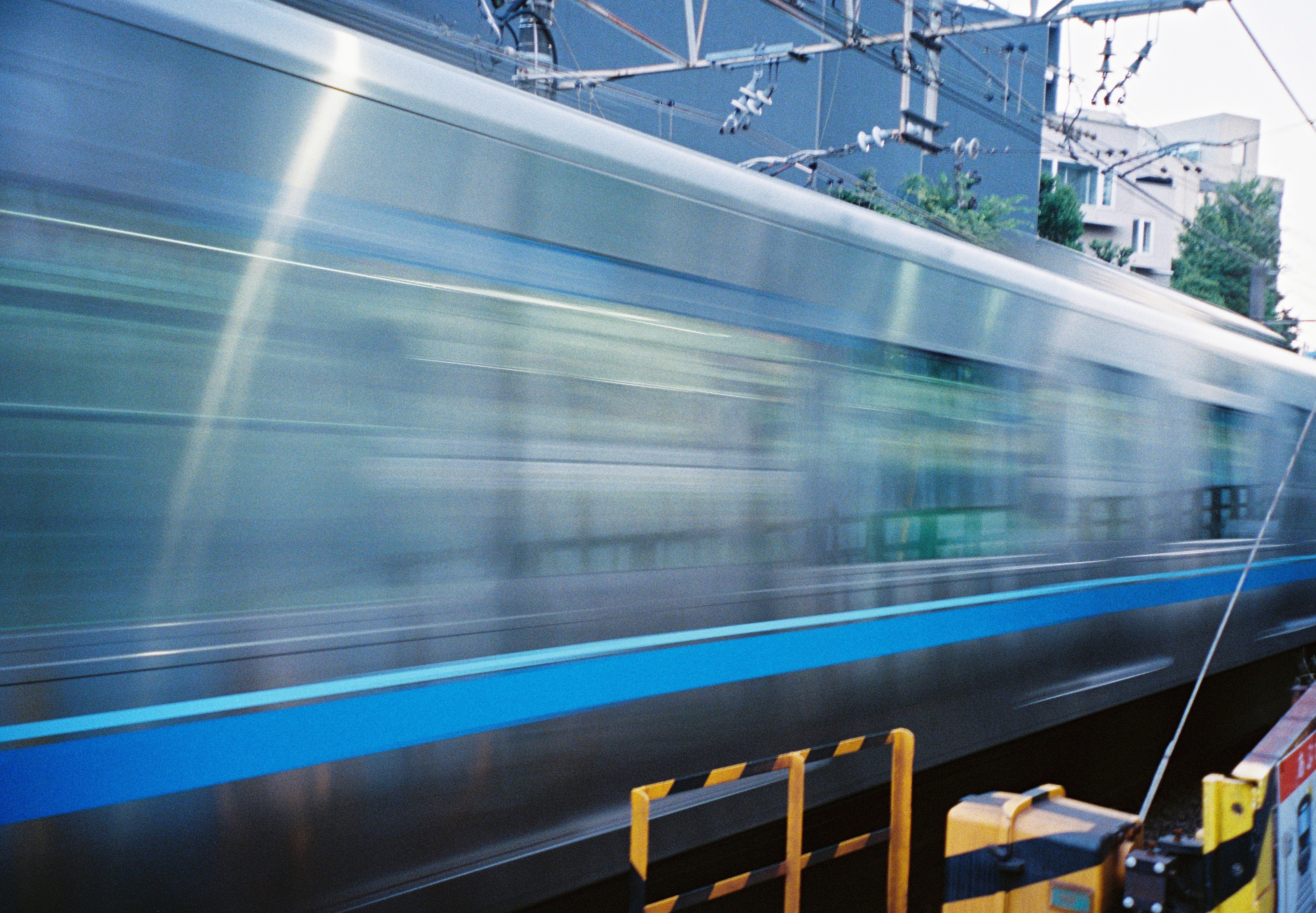 A blue-striped passenger train blurs across the frame beneath overhead railway wires and metal supports.
