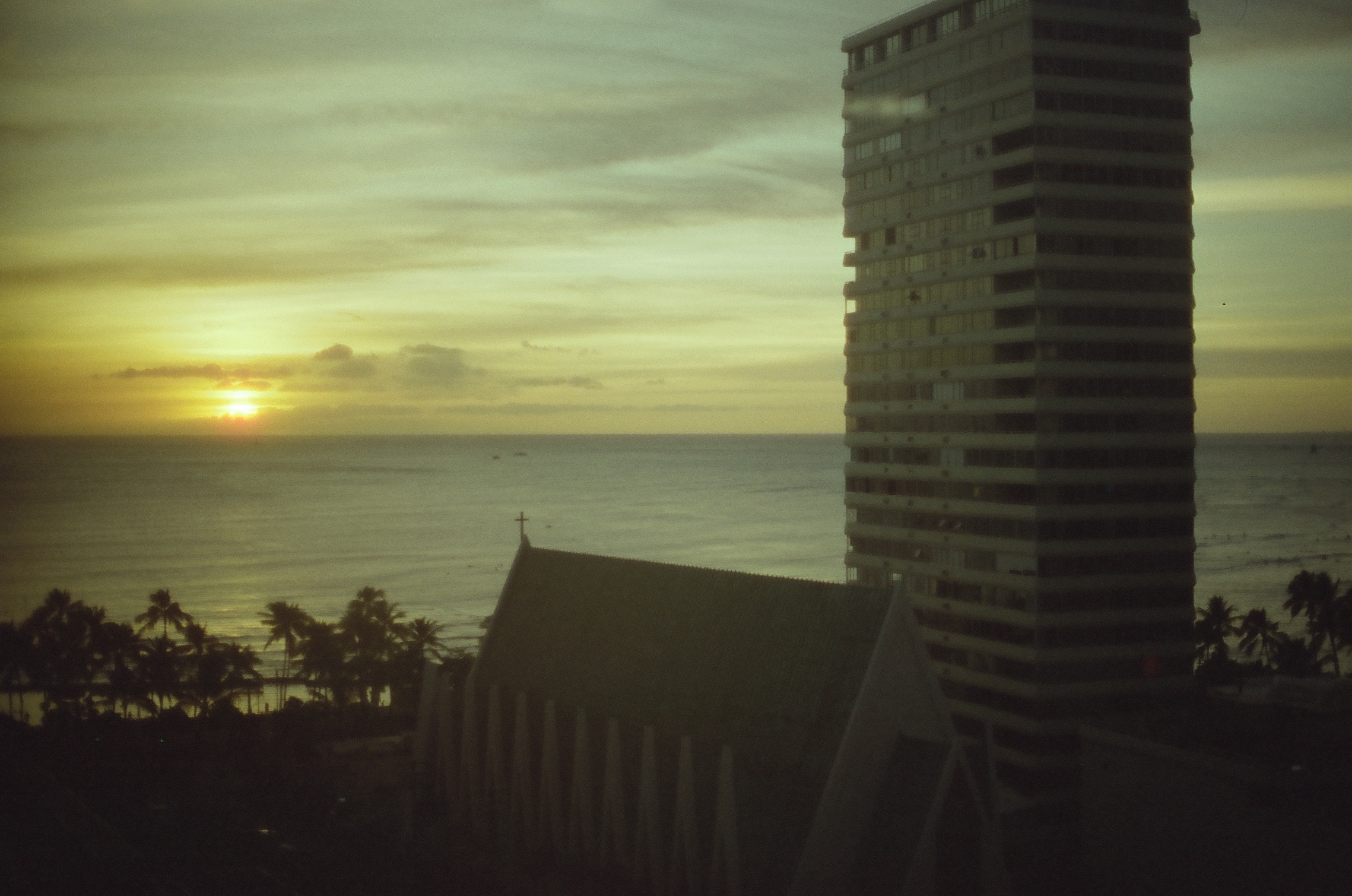 The low sun glows above calm water beside a tall building and dark sloping rooftops.