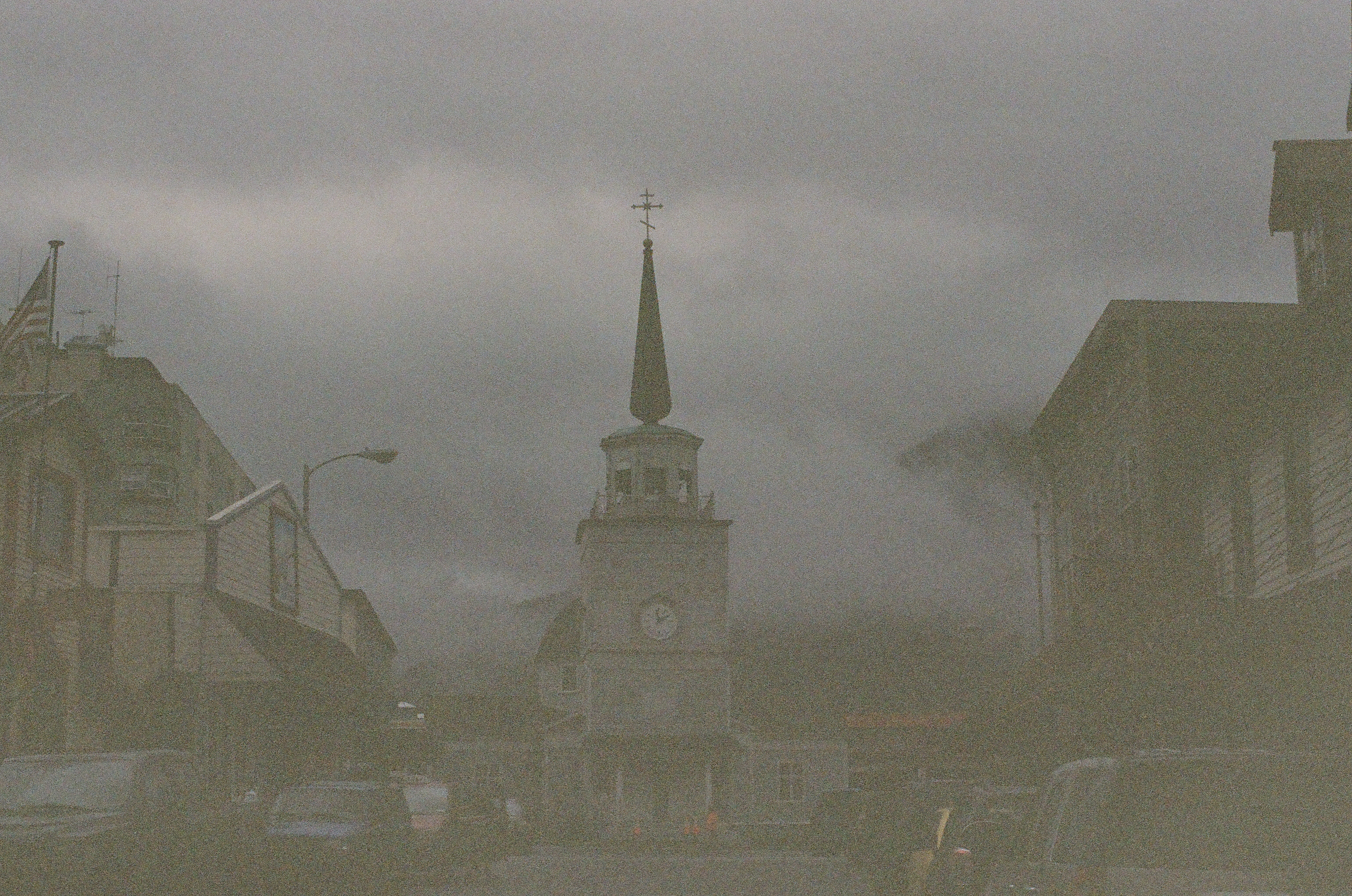 A pointed steeple rises at the end of a quiet street softened by gray rain or mist.