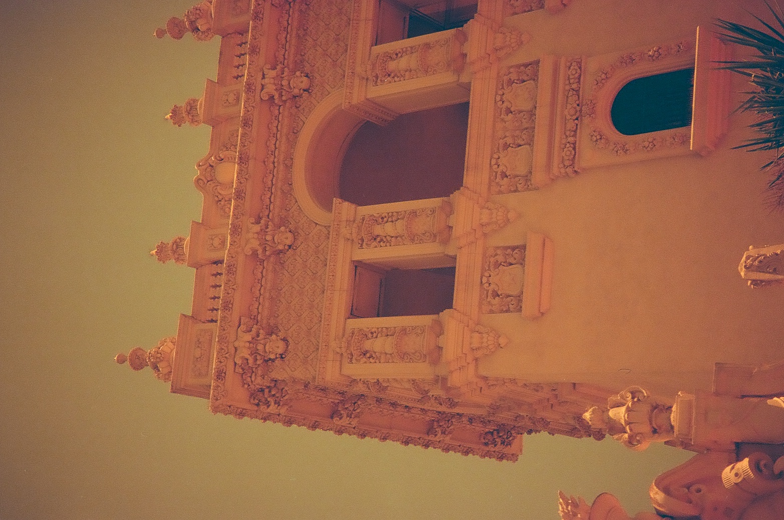 An ornately carved tower facade with balconies and a central arched opening rises against a pale green sky, framed by a palm frond.