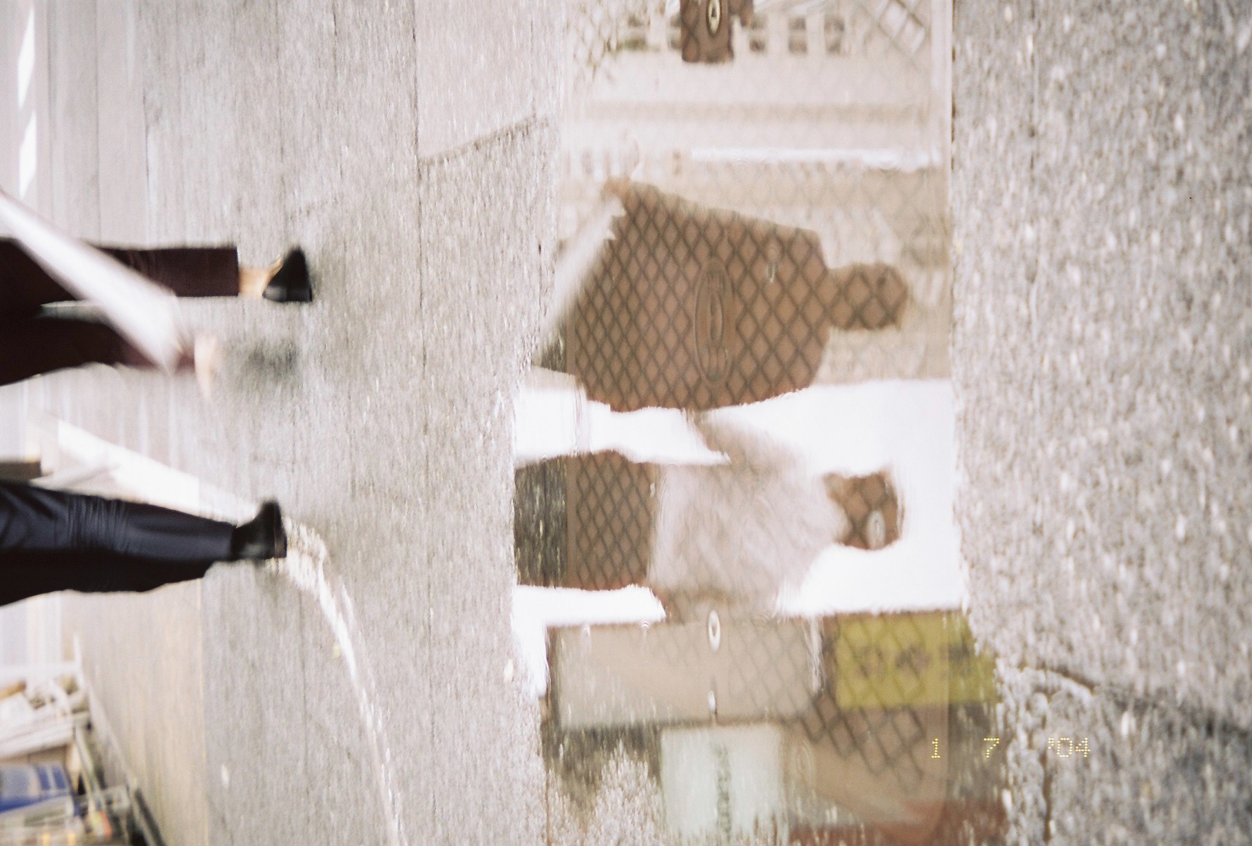 A puddle reflects two people walking side by side, one in dark clothing and one in a light shirt, with inverted pedestrians' legs along the lower edge.