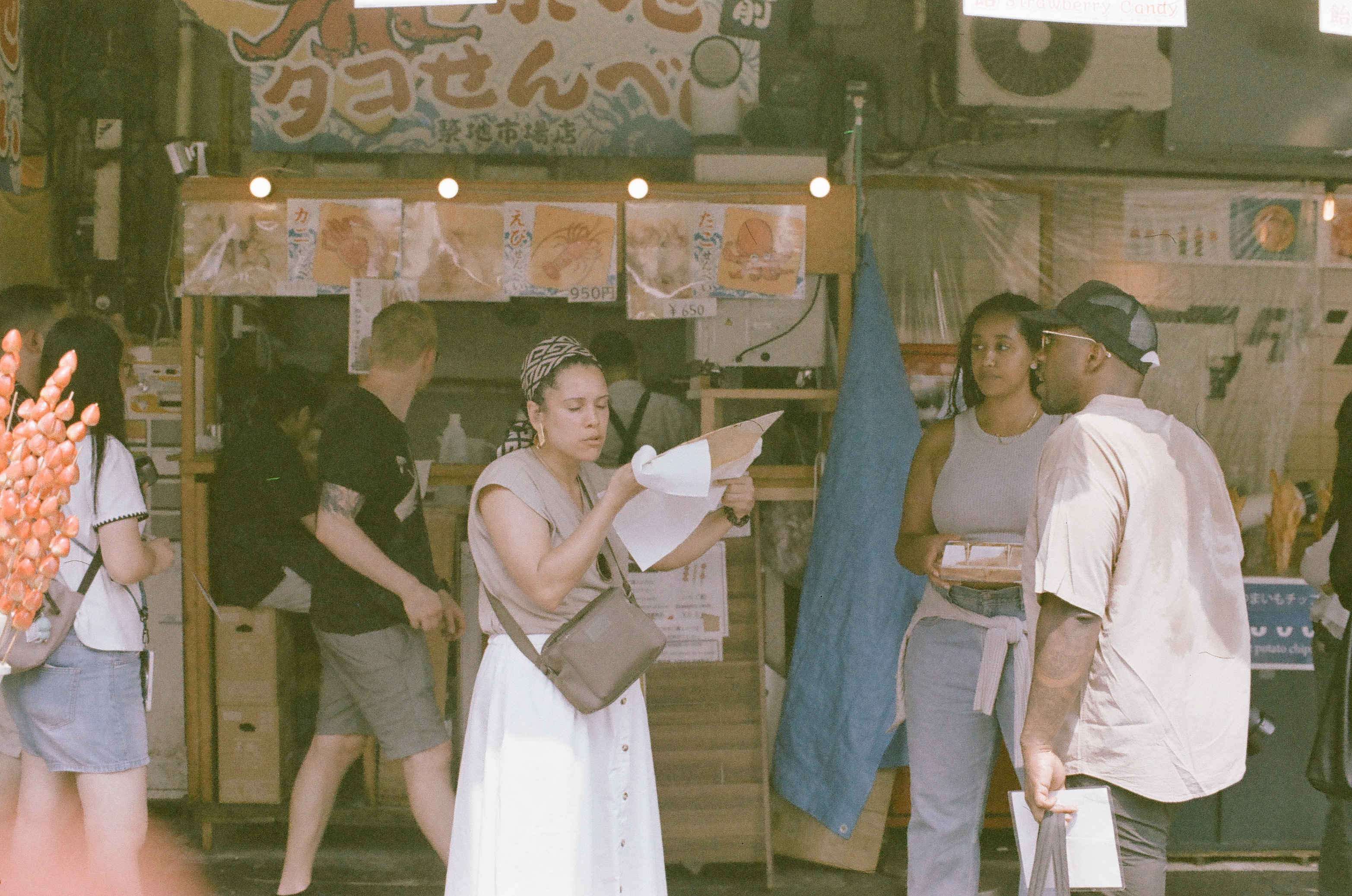 Lifting food from a folded paper wrapper, a woman pauses beneath a market stall banner while two other visitors stand nearby holding a takeout box.