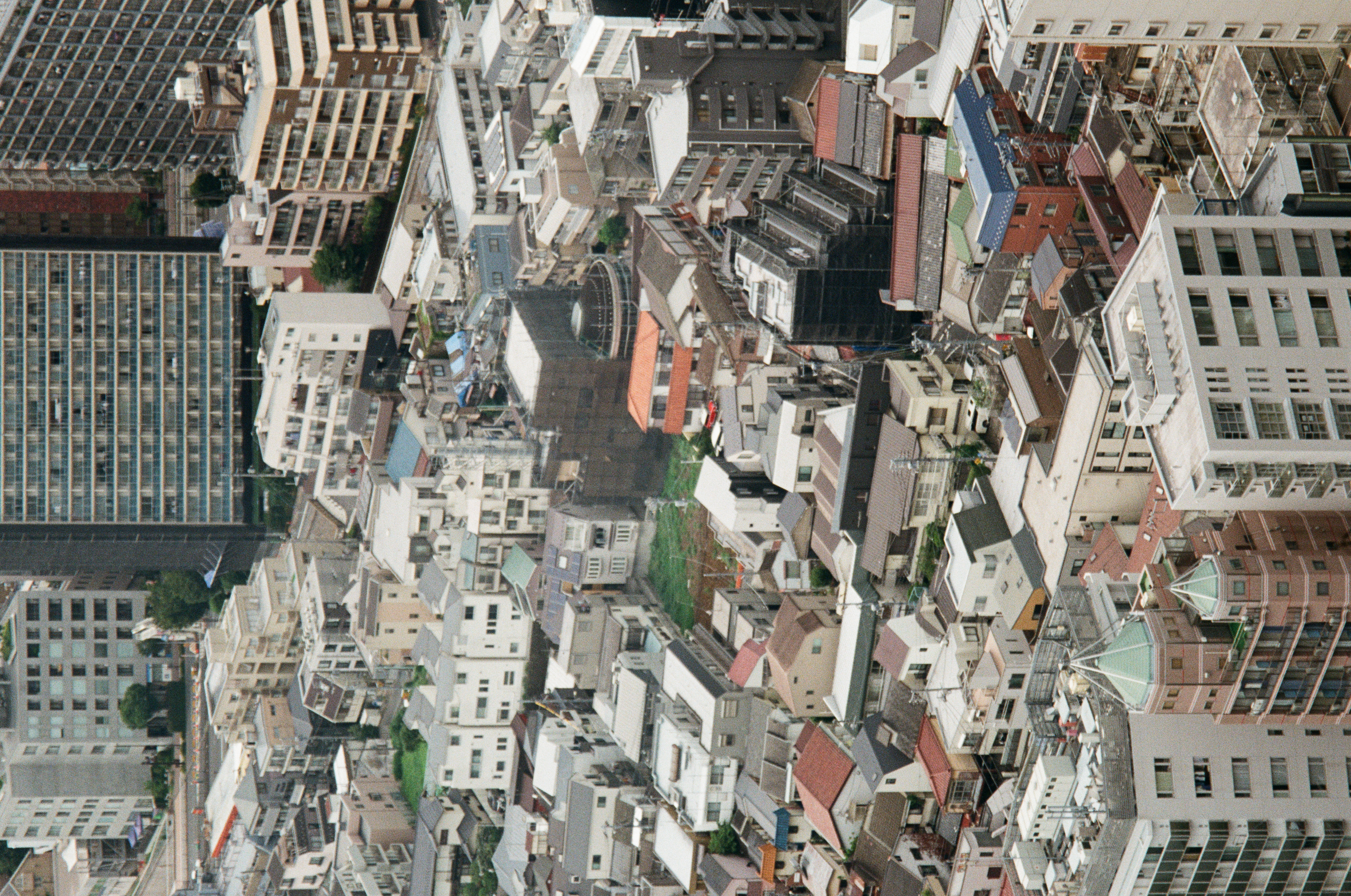Tiled house roofs, a laundry-railed apartment slab, and one vacant green lot crowd together in a tightly compressed rooftop view.