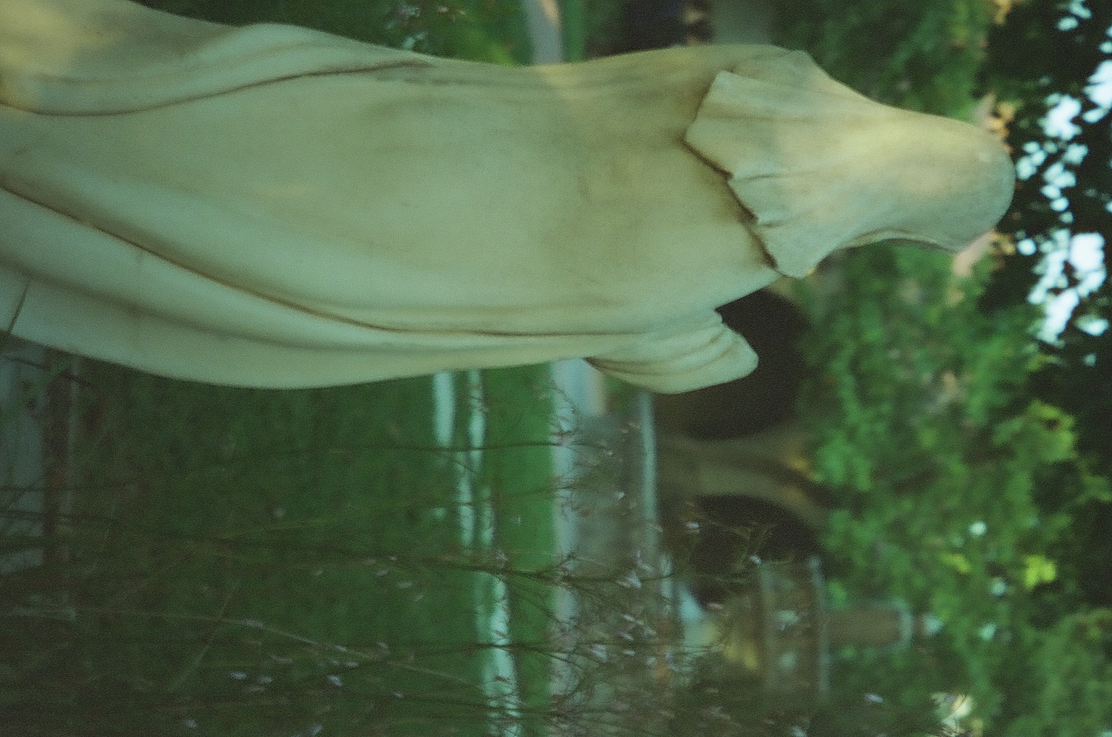 A pale robed statue stands among green trees and low fencing with one raised hand turned away from the viewer.