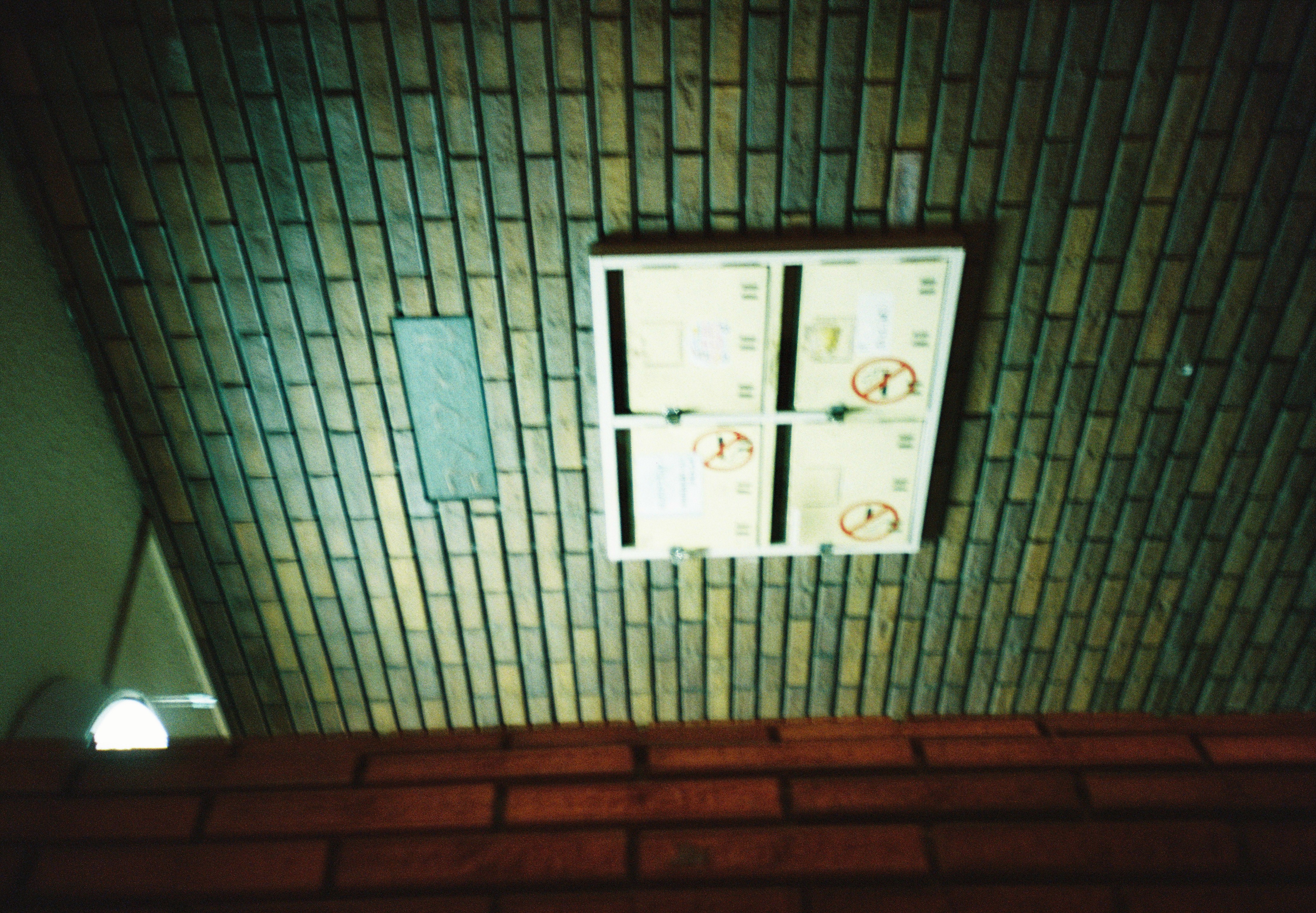 Four small metal mailboxes with taped doors hang on a green-tiled brick wall beneath a glowing ceiling lamp.