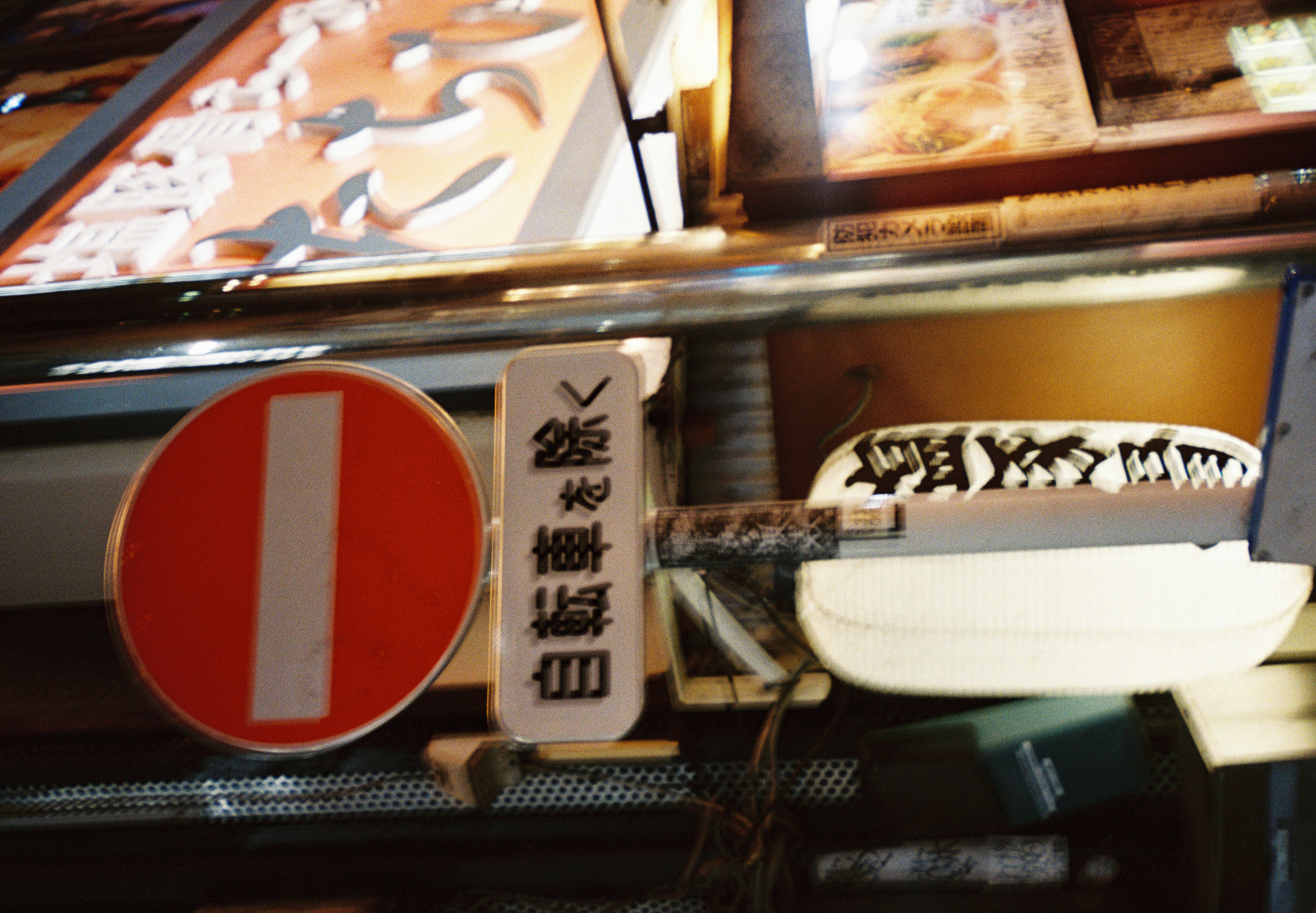 A red no-entry sign and smaller Japanese street signs stand before a brightly lit storefront at night.