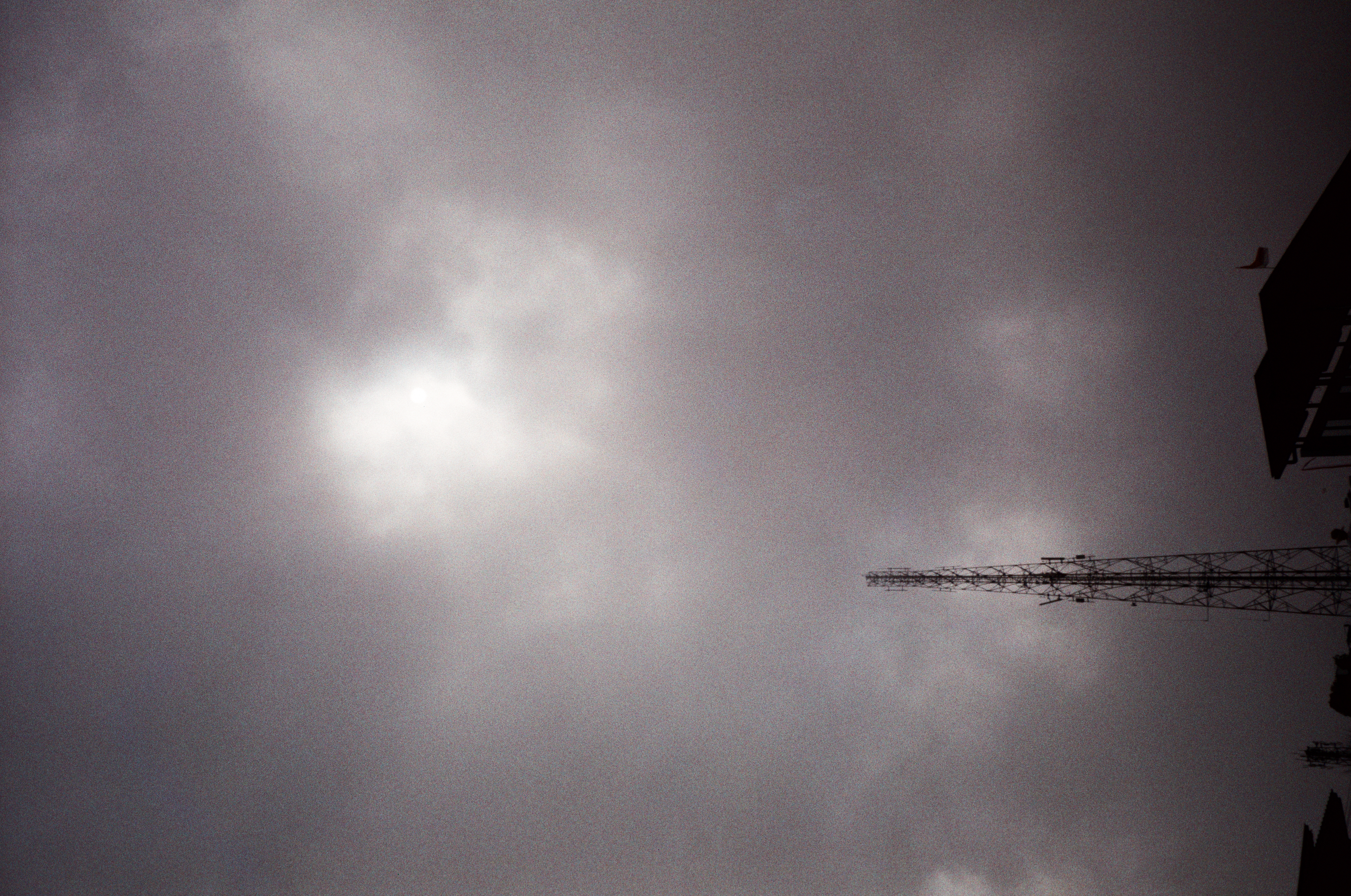 A lattice tower and dark roof silhouettes sit below a diffuse bright patch within an otherwise gray, clouded sky.