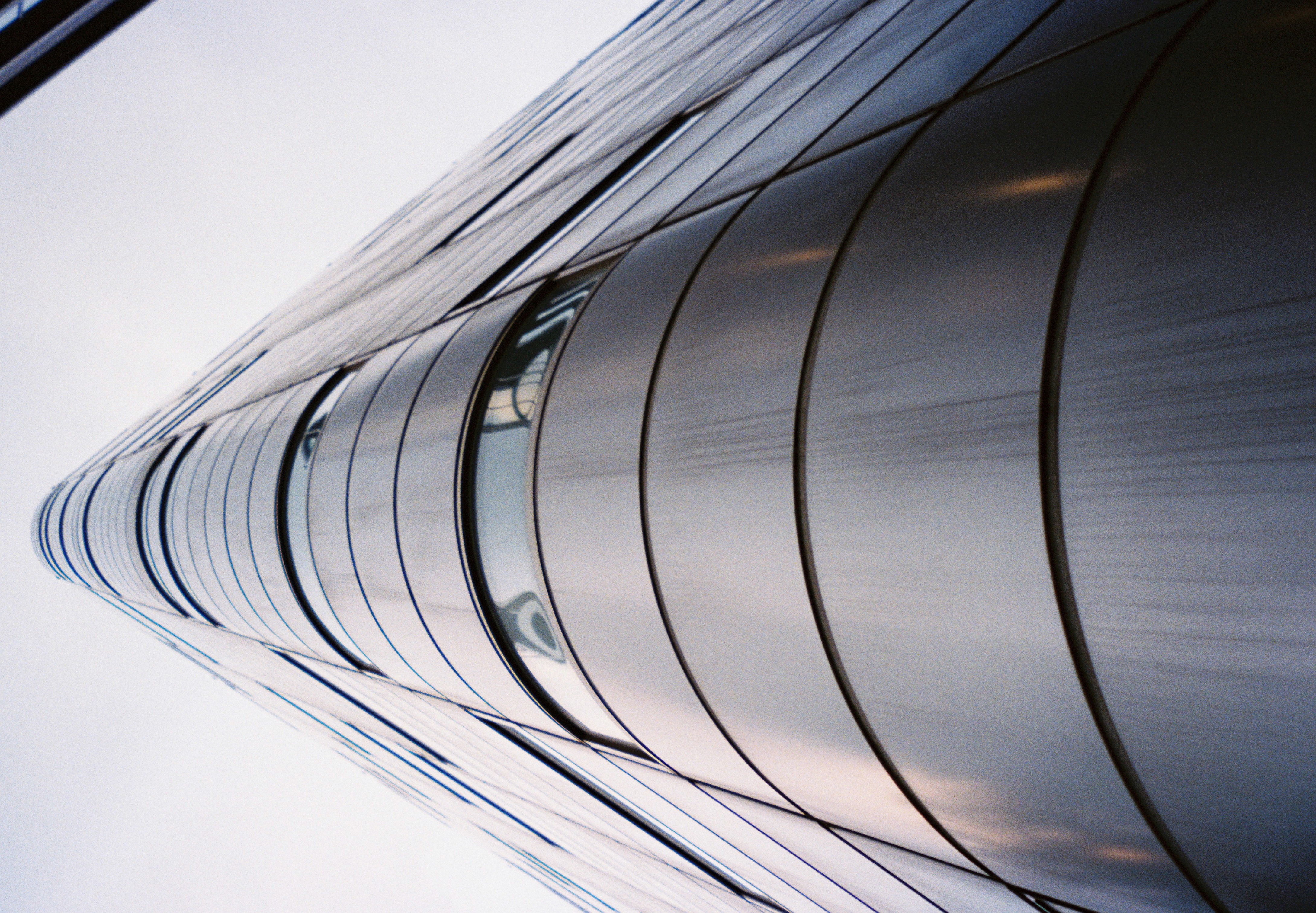 The rounded corner of a metal-clad building rises from the bottom edge toward a pale sky, its horizontal bands and window strips converging overhead.