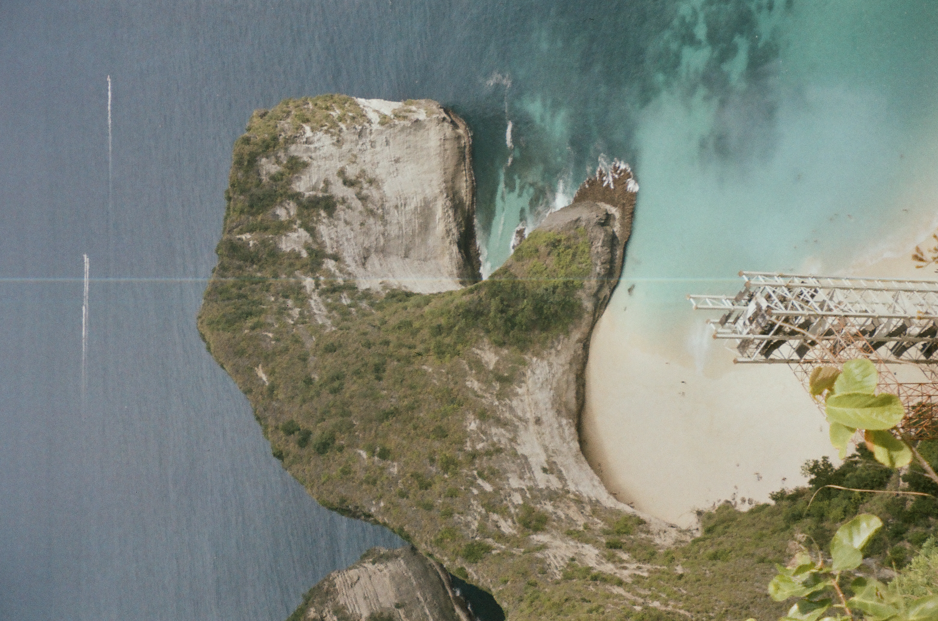 A rugged green headland encloses turquoise shallows beneath a broad field of calm gray-blue water.