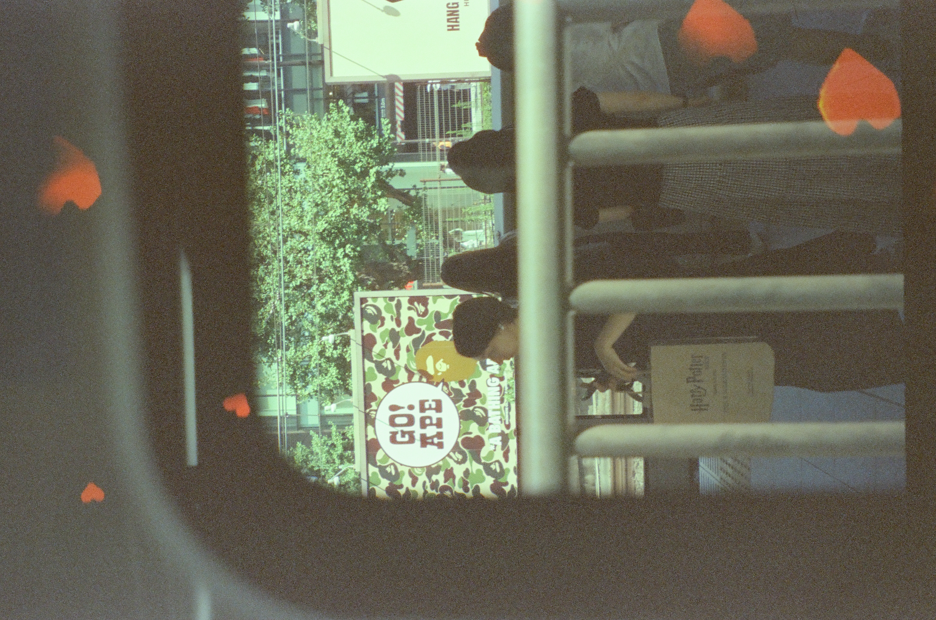 Pedestrians stand at a metal railing beneath a camouflage-patterned billboard reading 'GO! APE,' one carrying a bag printed 'Harry Potter,' with orange heart shapes in the foreground.