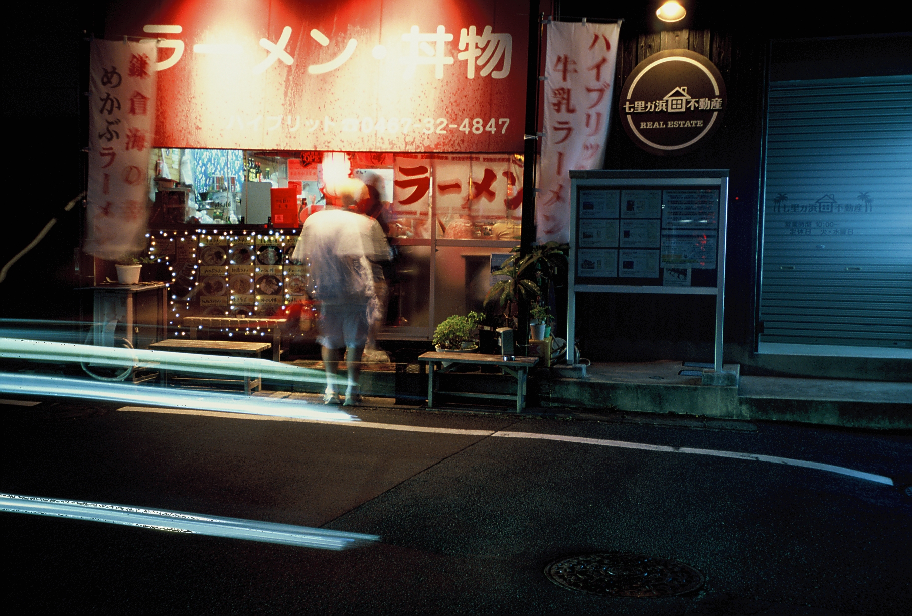 Long exposure light trails streak across a road as a blurred figure stands before a ramen counter lit with red signage and string lights.