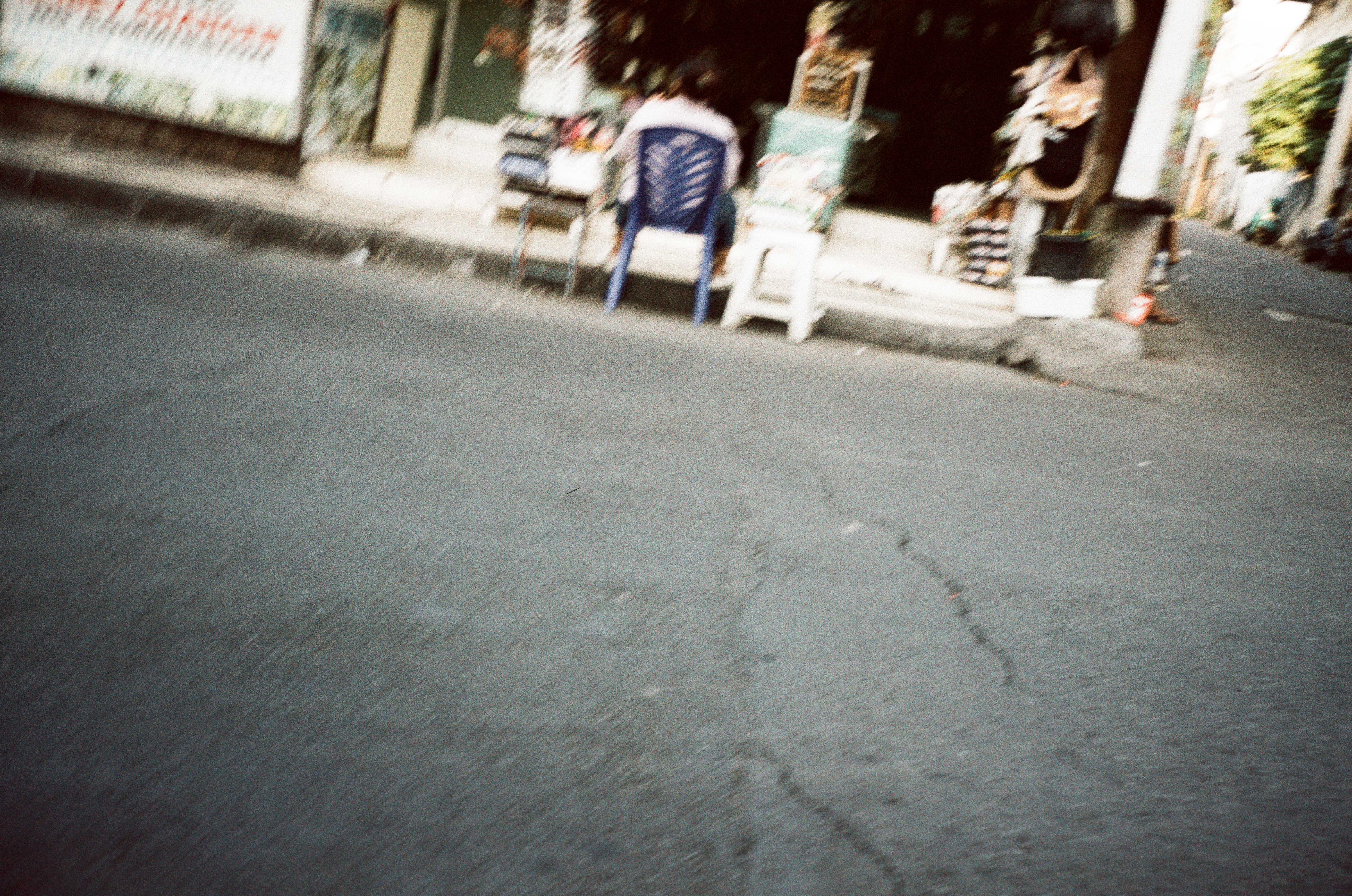 Cracked asphalt fills a tilted frame, rising toward a person seated on a blue plastic chair beside a white stool at a roadside stall.