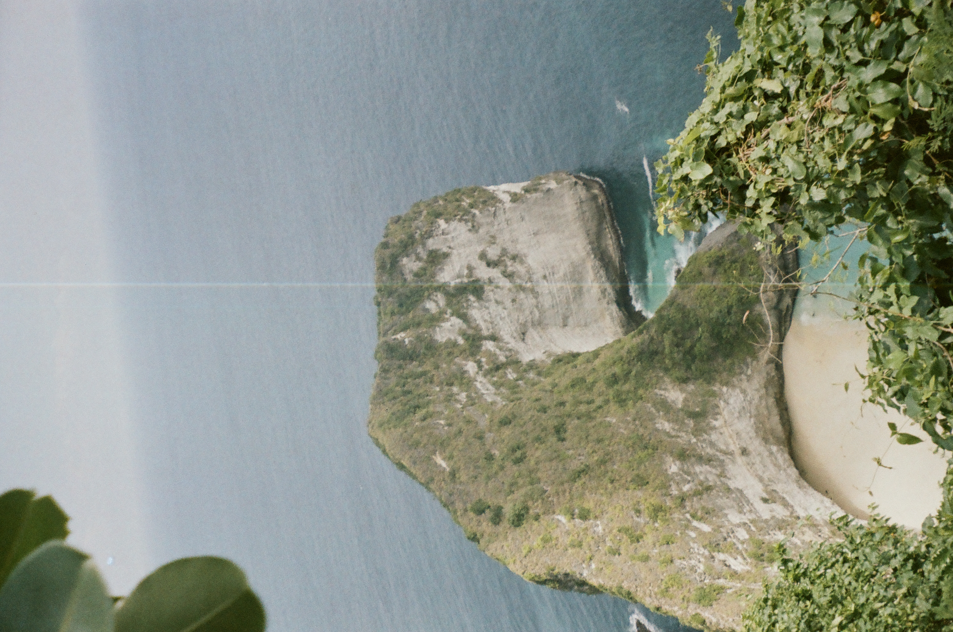 A green and pale rock formation descends toward a sandy cove amid broad blue water, framed by leafy vegetation above and below.