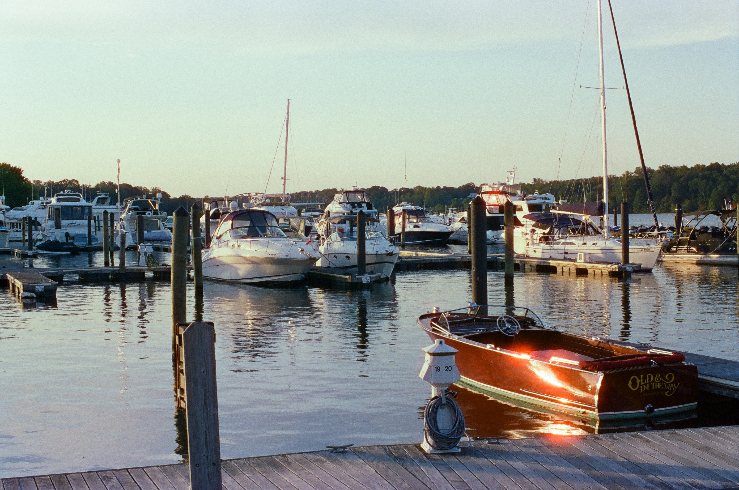 The same wooden runabout and marina appear again under cooler, brighter light, with more boats and a taller sailboat mast visible.