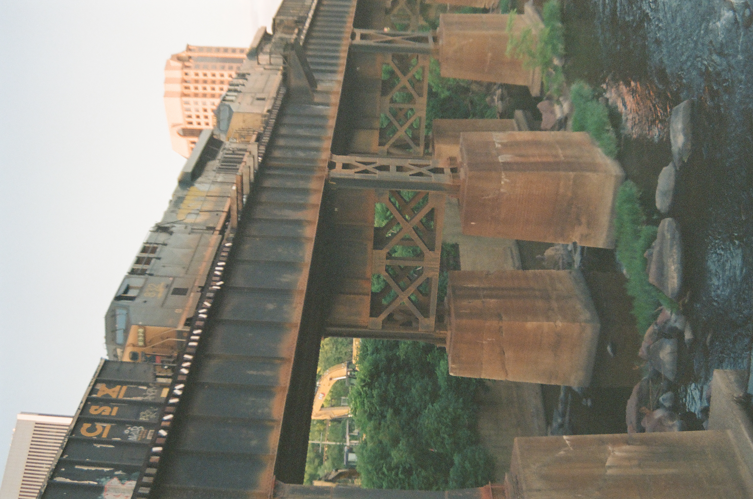 A freight train marked 'CSX' crosses a rusted steel trestle above the river, with a tall building lit by evening light behind it.