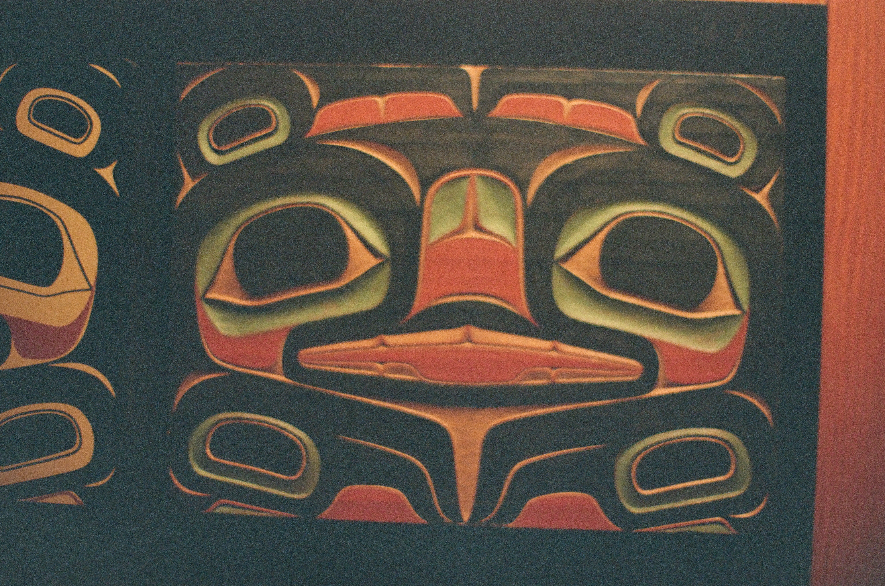 Red, black, blue, and cream shapes form a symmetrical carved face across a flat wooden panel.