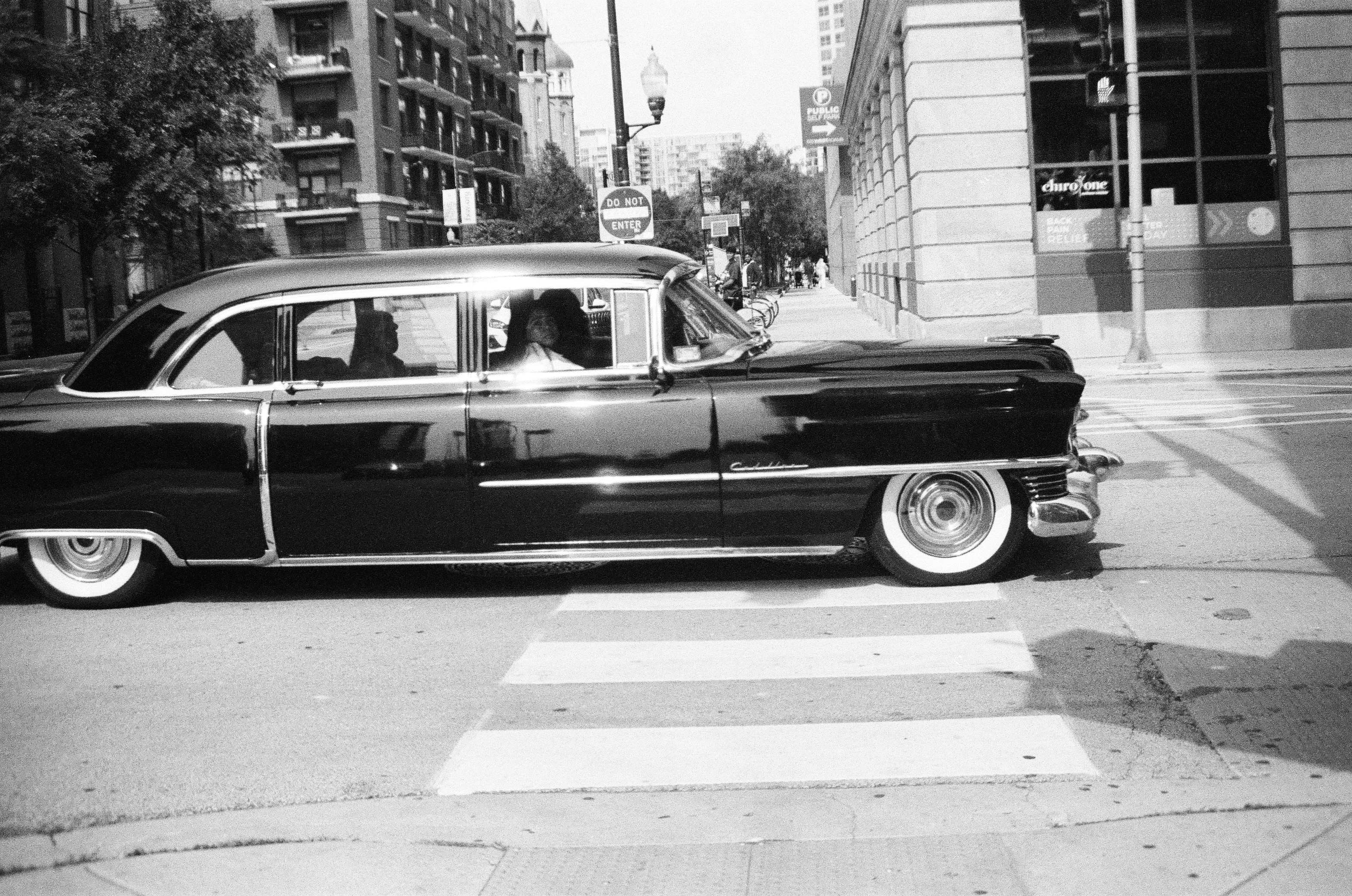 A long dark vintage car crosses a striped intersection while two occupants remain visible through its side windows.