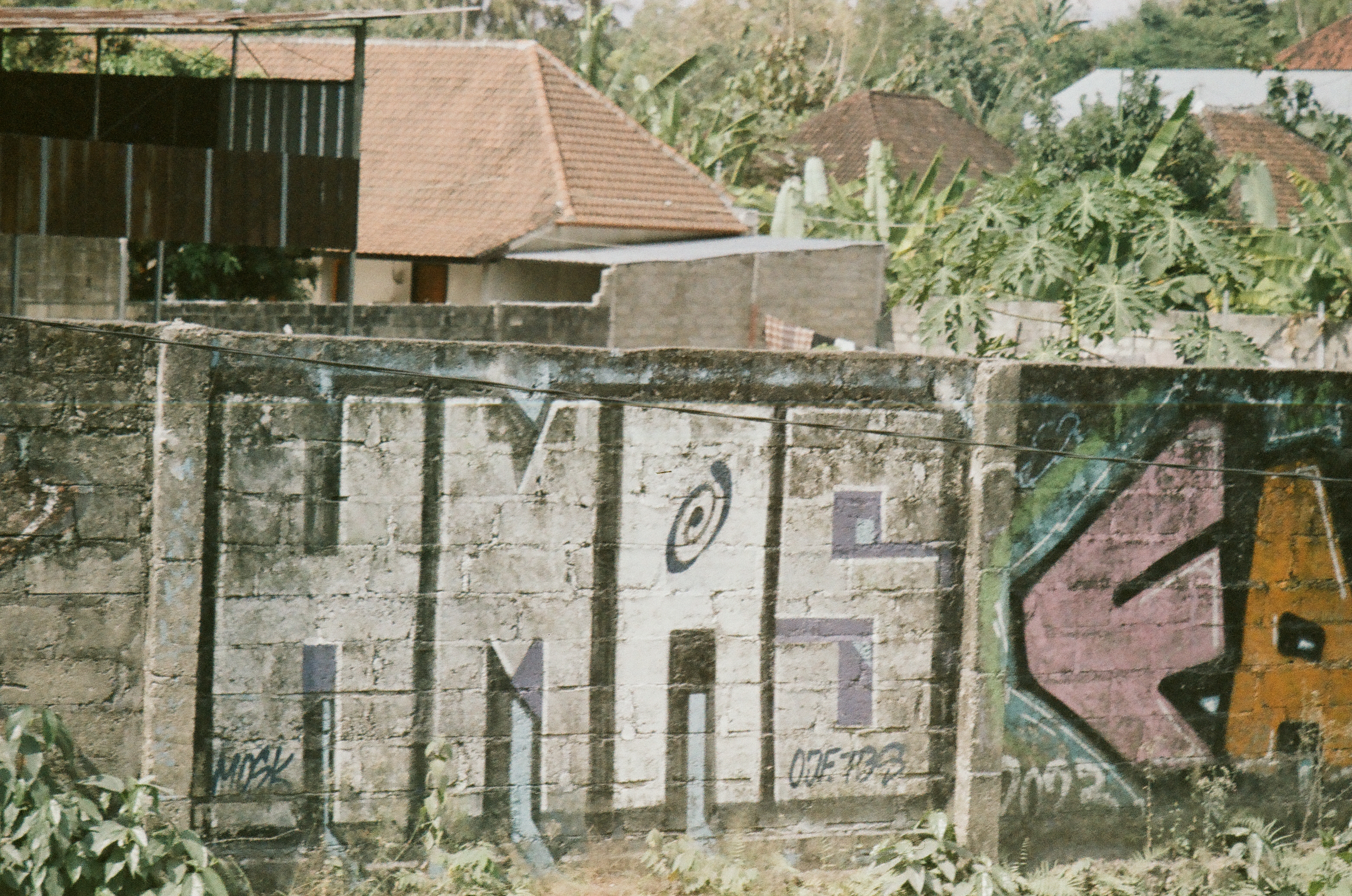 Large painted letters and angular color blocks cover a weathered wall beneath tiled roofs and trees.
