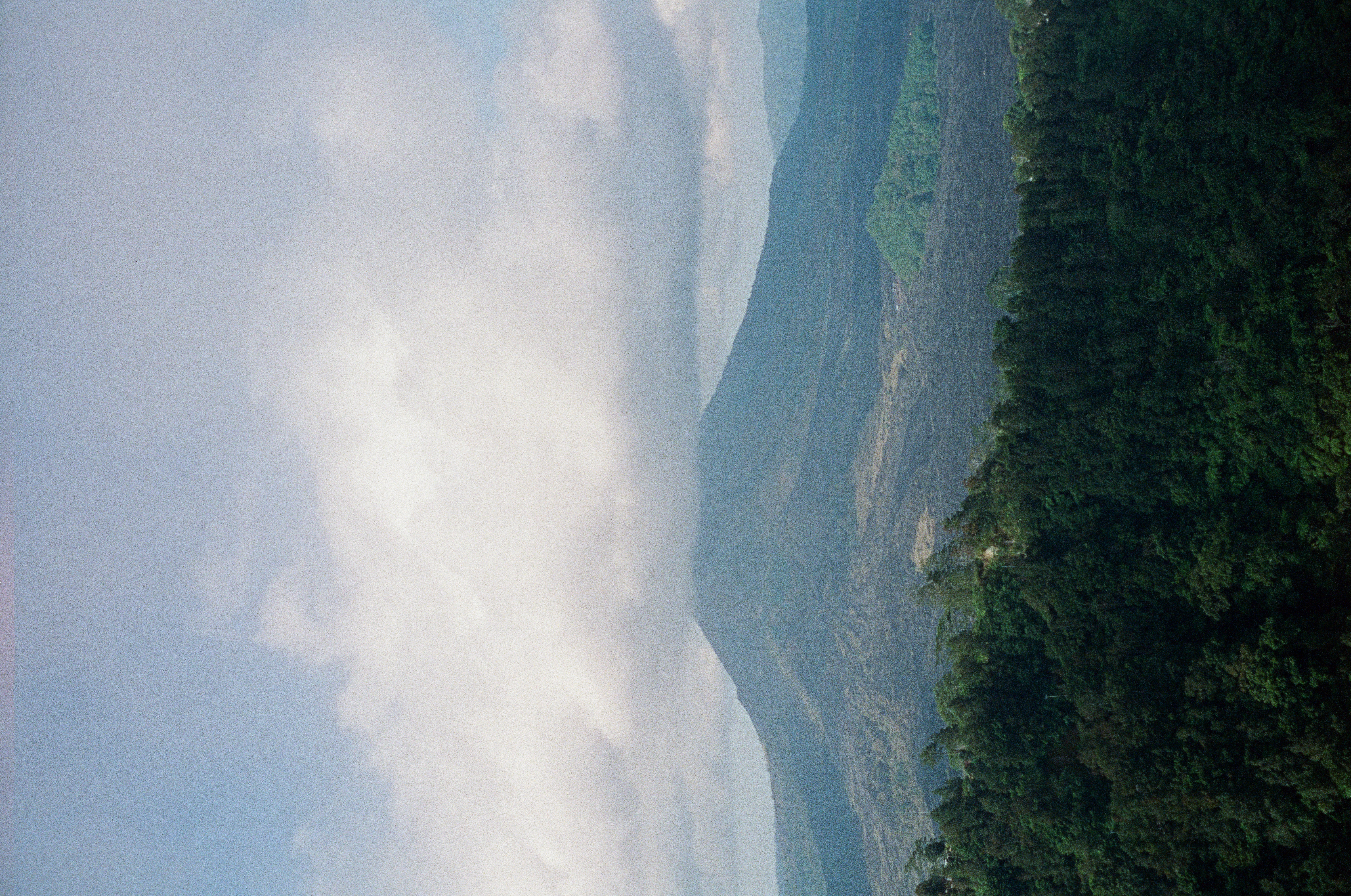A conical mountain with dark, flow-like bare slopes rises beyond a forested ridge, its summit hidden under a thick bank of white cloud.