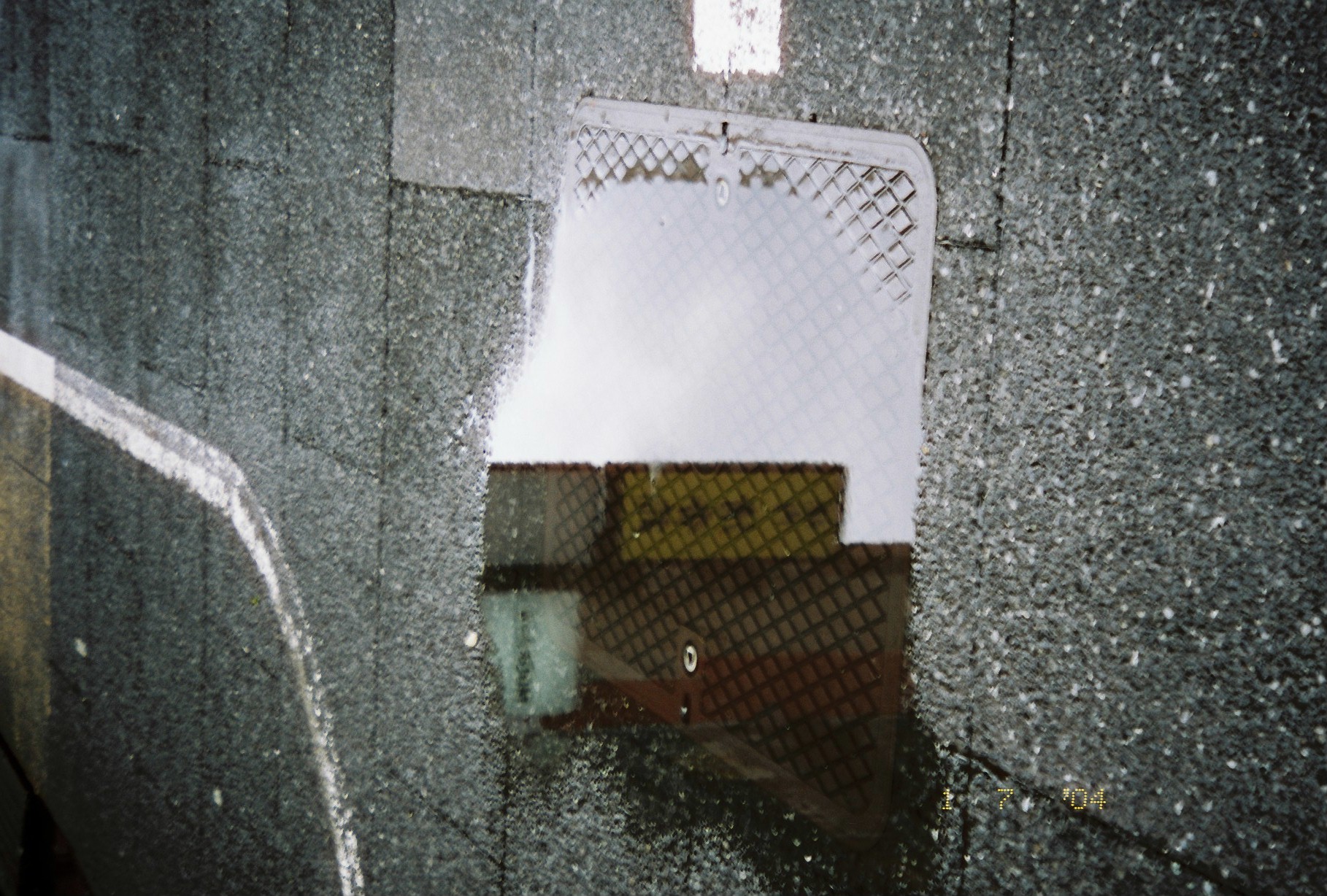 A sharp-edged puddle on dark block paving reflects white sky on its left half and a yellow-lit panel beside a red-brown gridded facade on its right.