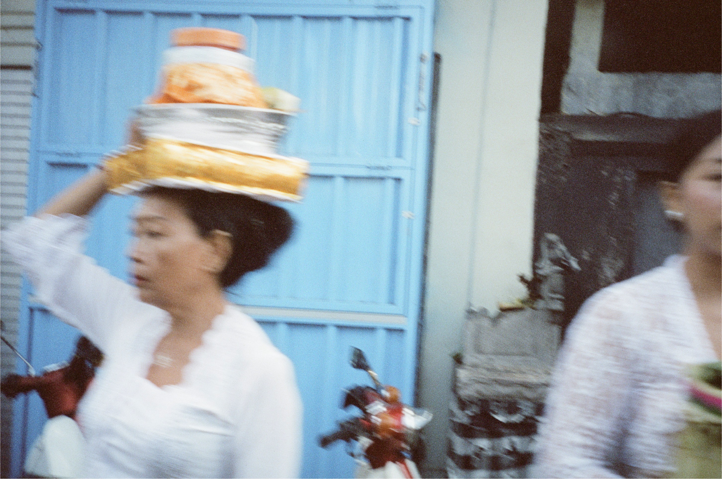 An adult in white steadies a gold-and-orange tiered load on their head before a bright blue metal gate.